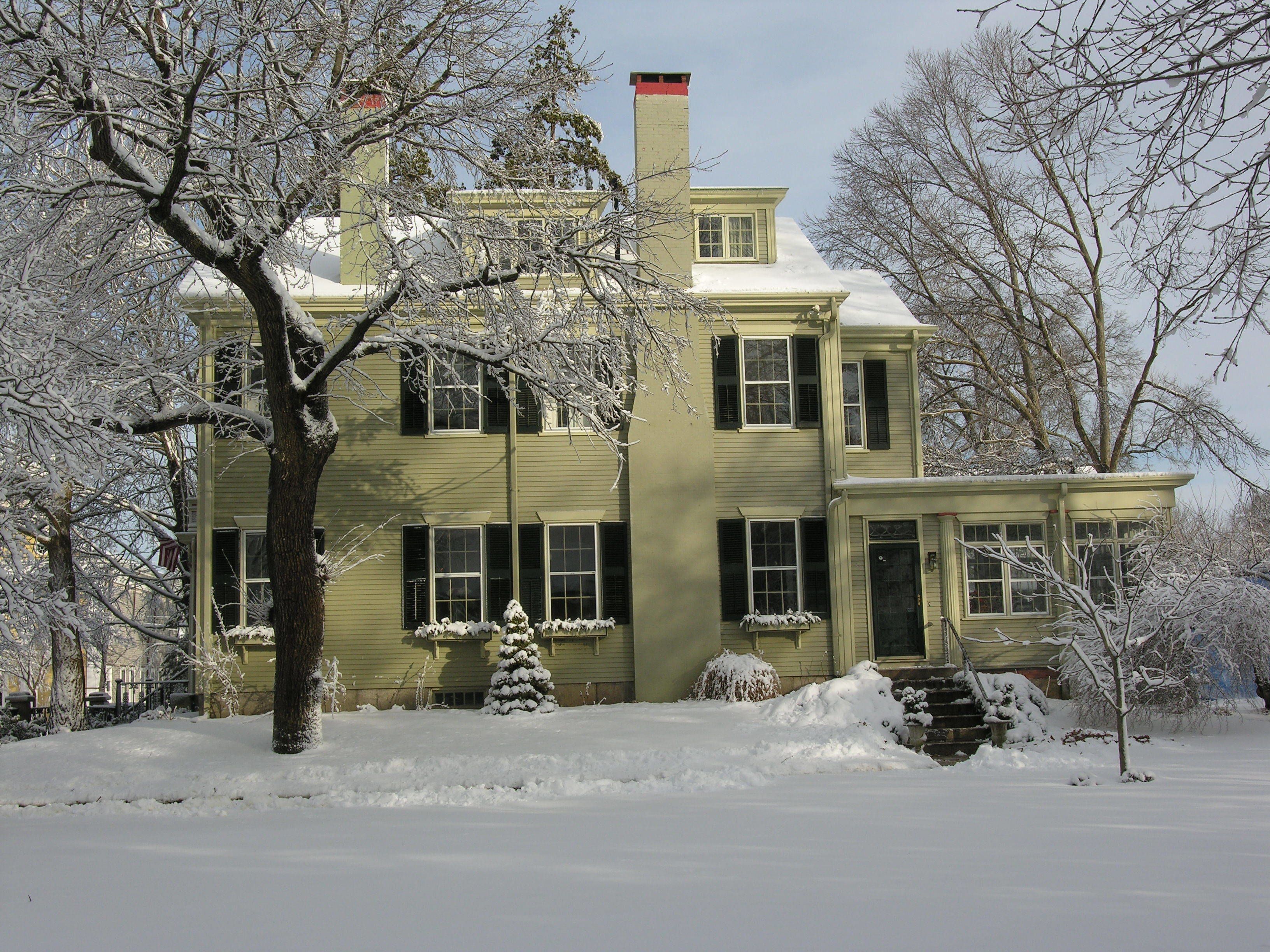 A two-story house with a chimney, covered in snow, surrounded by bare trees.