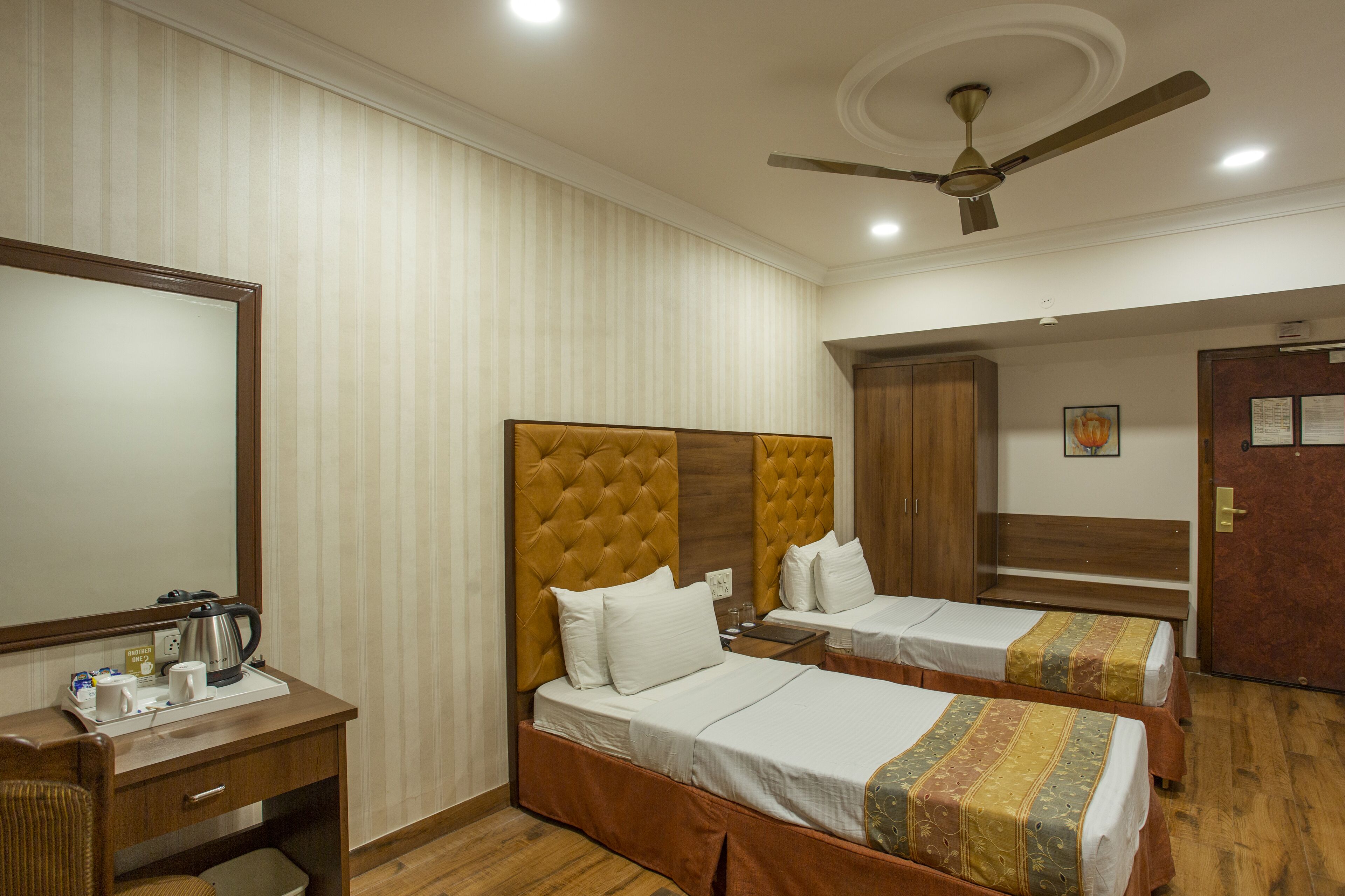 A hotel room with two single beds, a desk with a mirror, a coffee maker, and a ceiling fan.