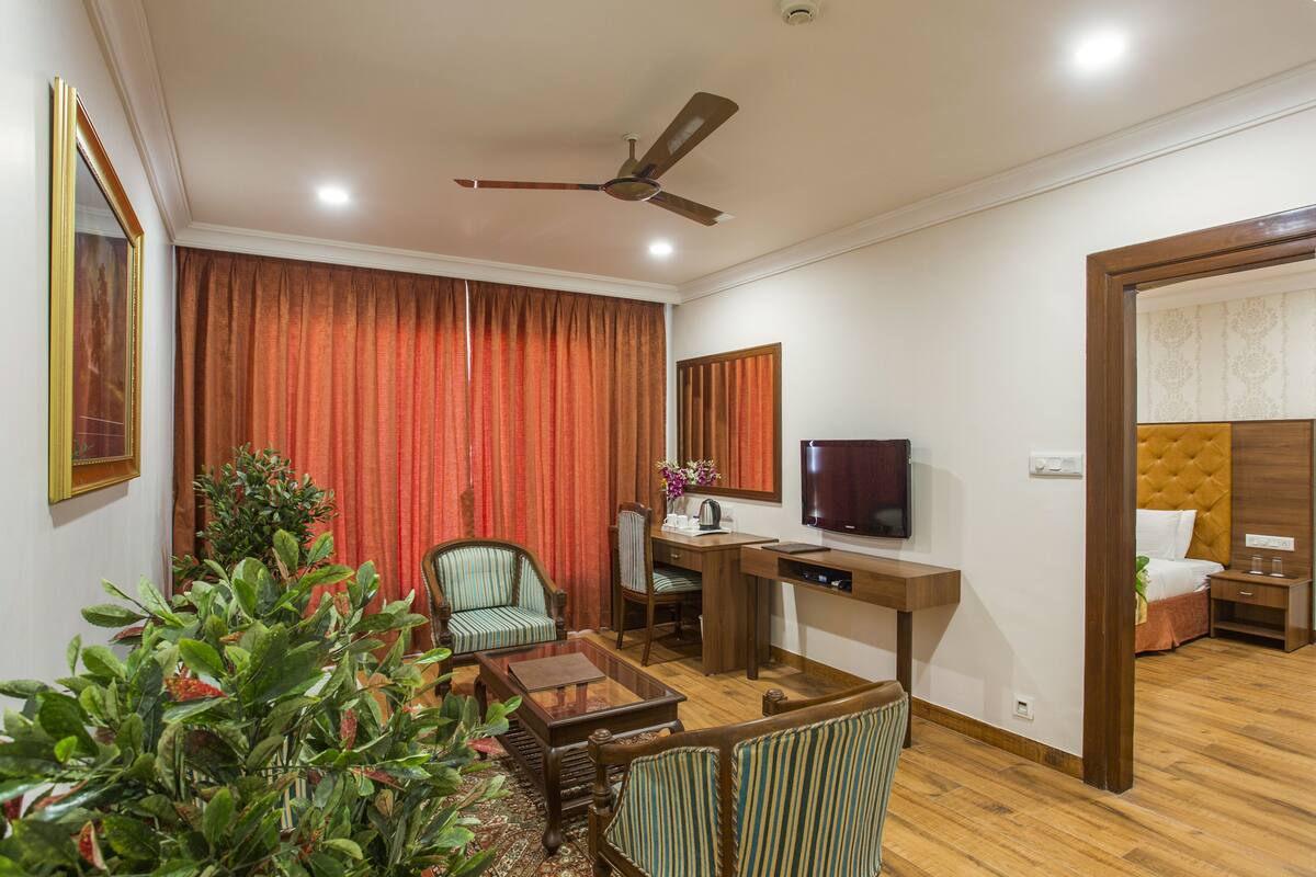 Une chambre d’hôtel avec un bureau en bois, une télévision, un lit doté d’une tête de lit capitonnée, un coin salon avec des fauteuils et une grande fenêtre ornée de rideaux rouges.