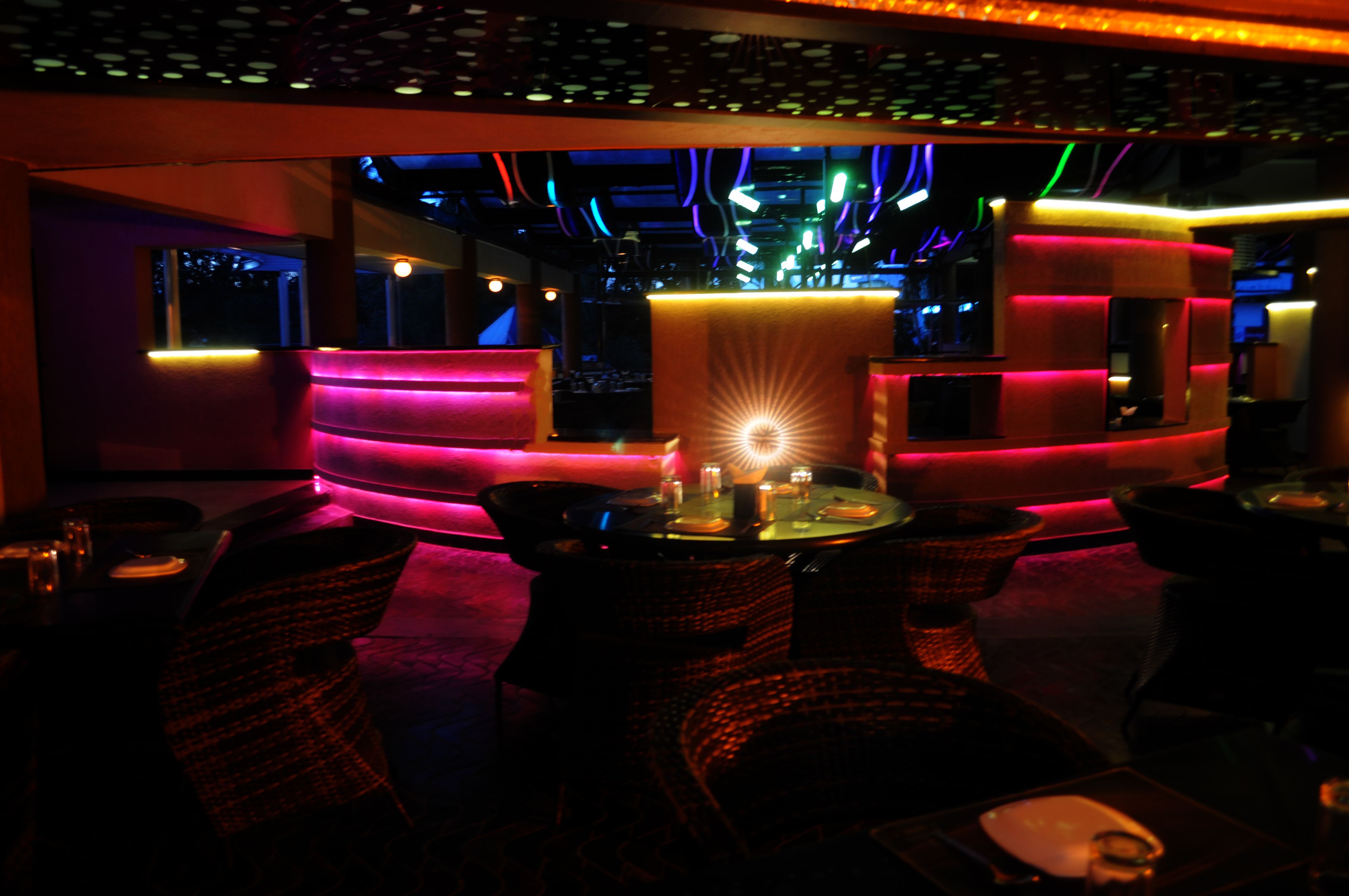 A dimly lit lounge area with a central table surrounded by wicker chairs, a neon-lit reception desk, and a view of the interior space.