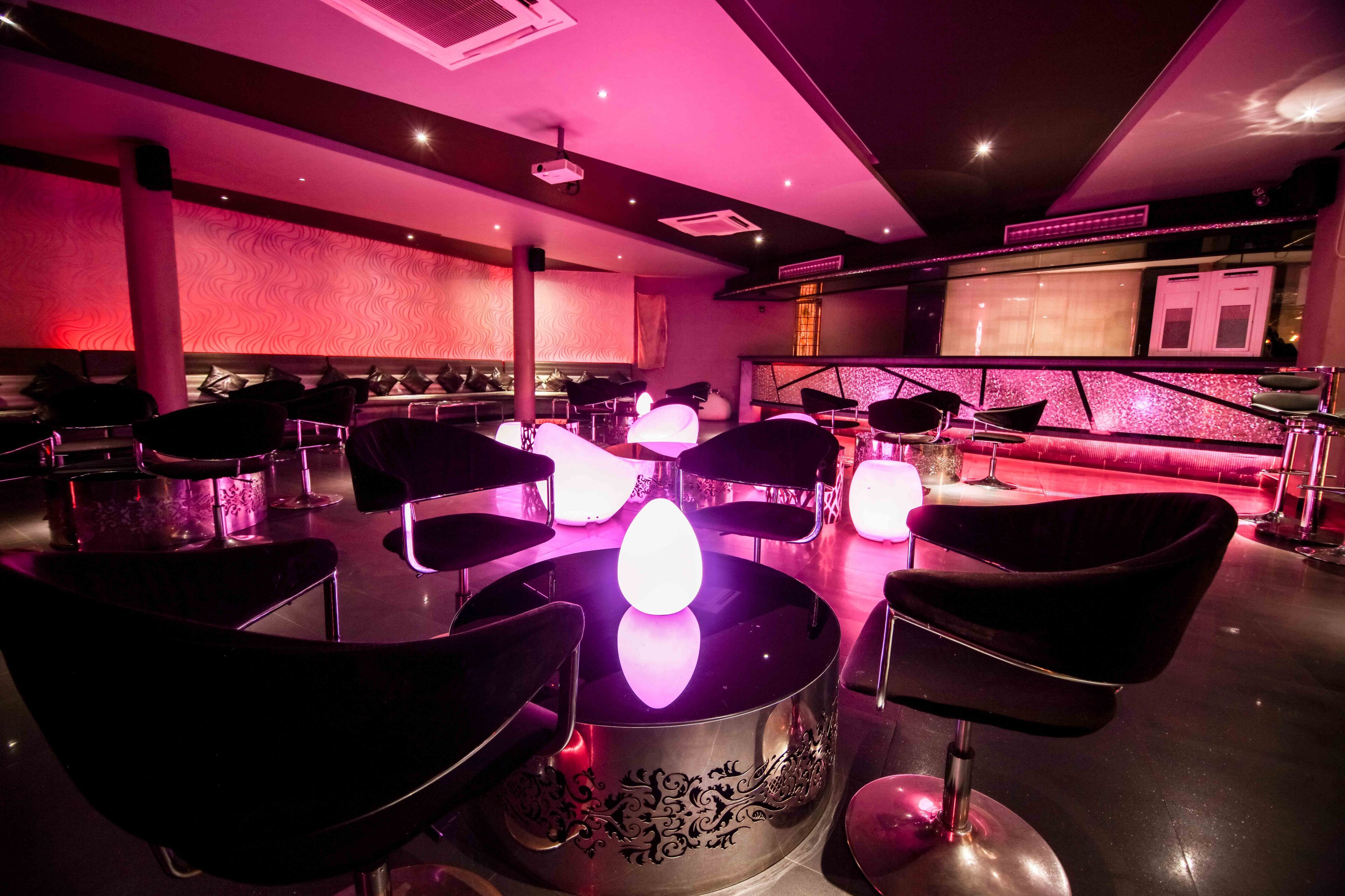Zona de bar con sillas negras modernas, una mesa central con luz, y una pared iluminada en rosa.