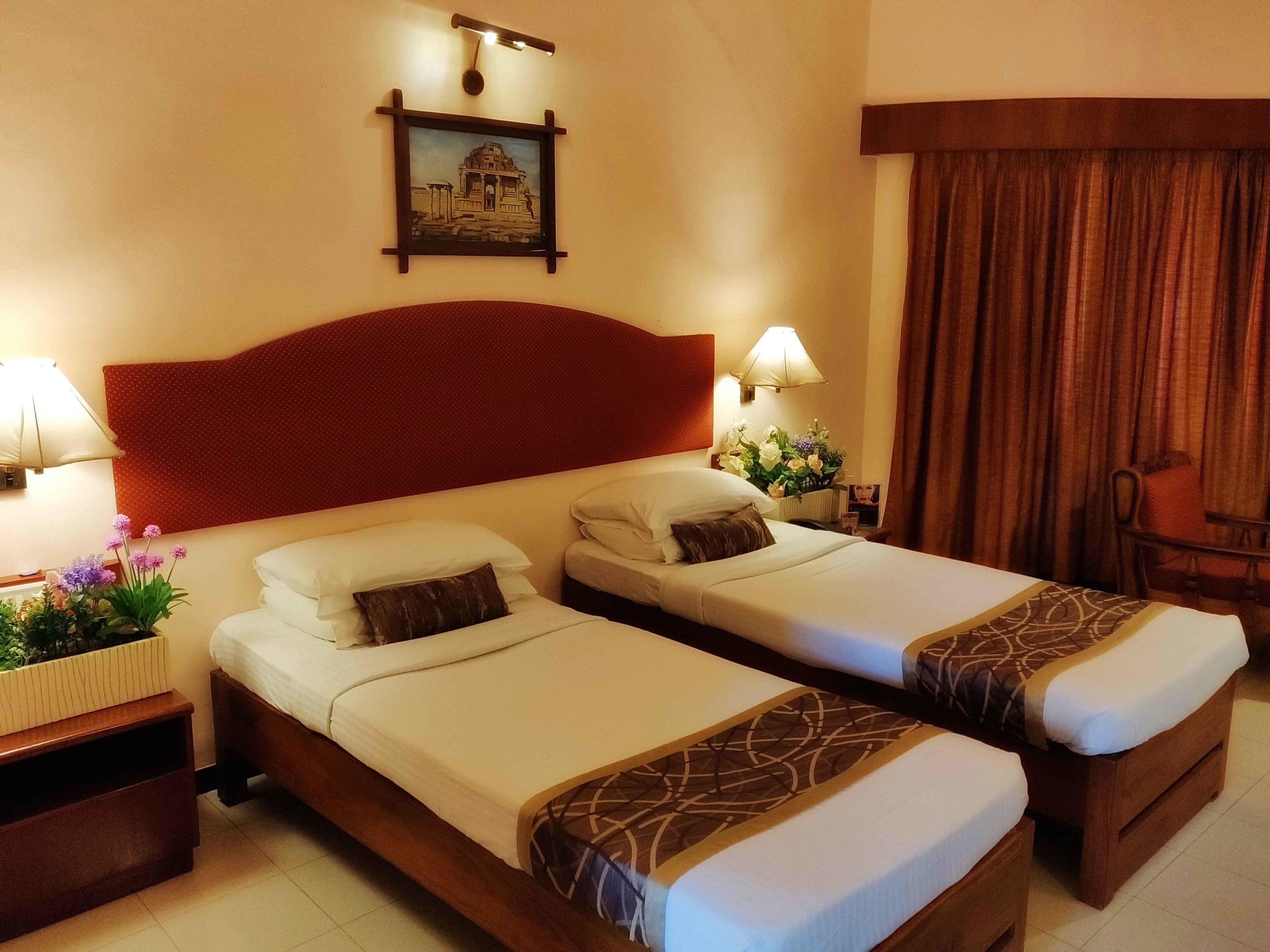 A hotel room with two single beds, a red headboard, a framed picture on the wall, a nightstand with a lamp, and a window with curtains.