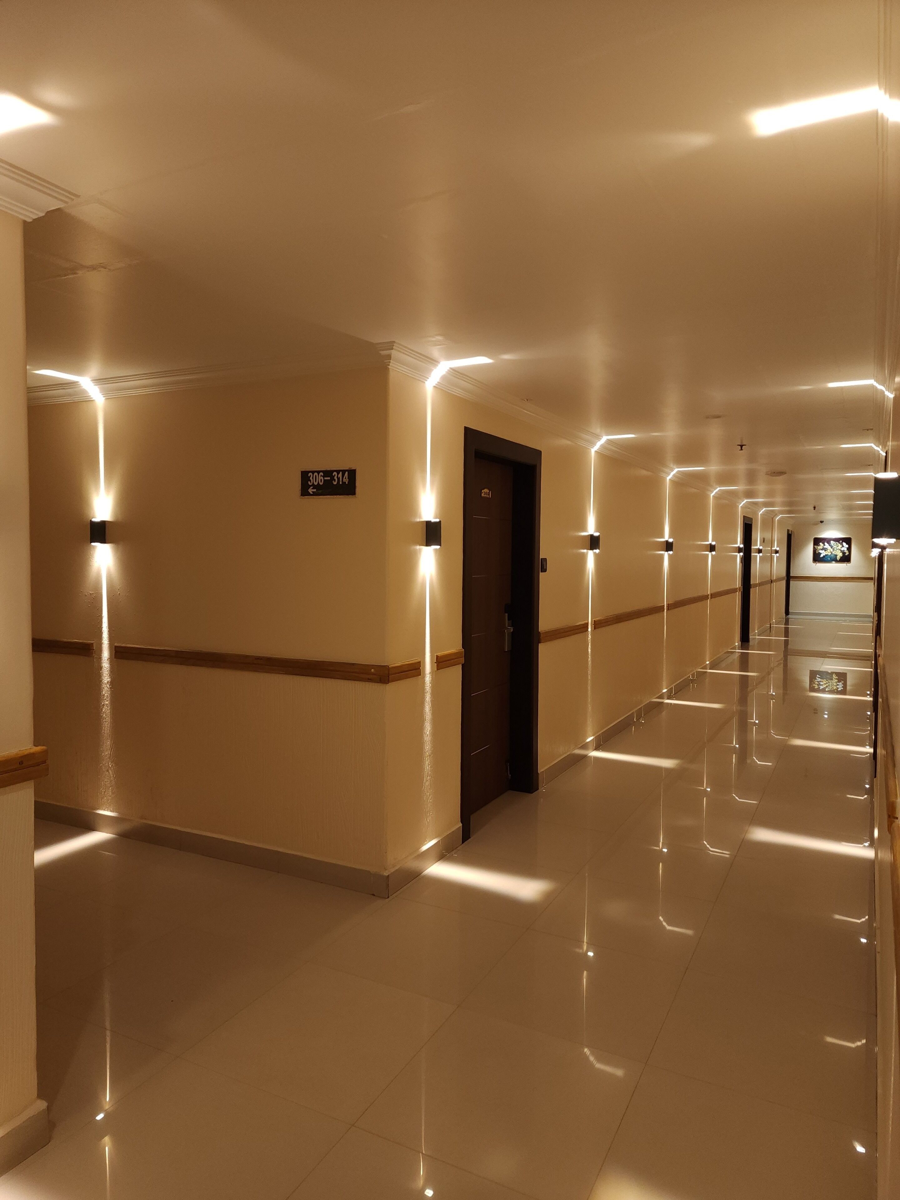 A hotel corridor with a polished floor, wall-mounted light fixtures, and a door with a sign indicating room numbers.
