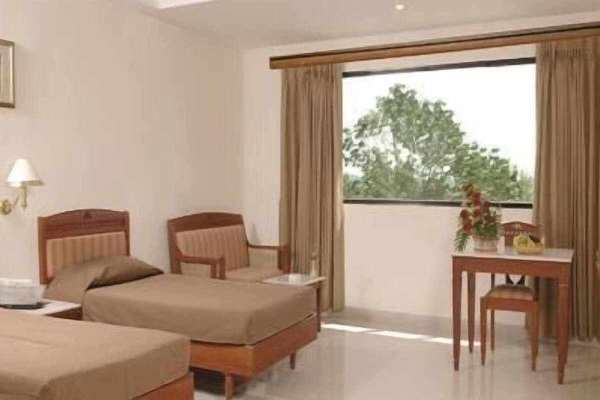 A hotel room with two beds, a desk, a chair, a television, and a window with a view of trees.