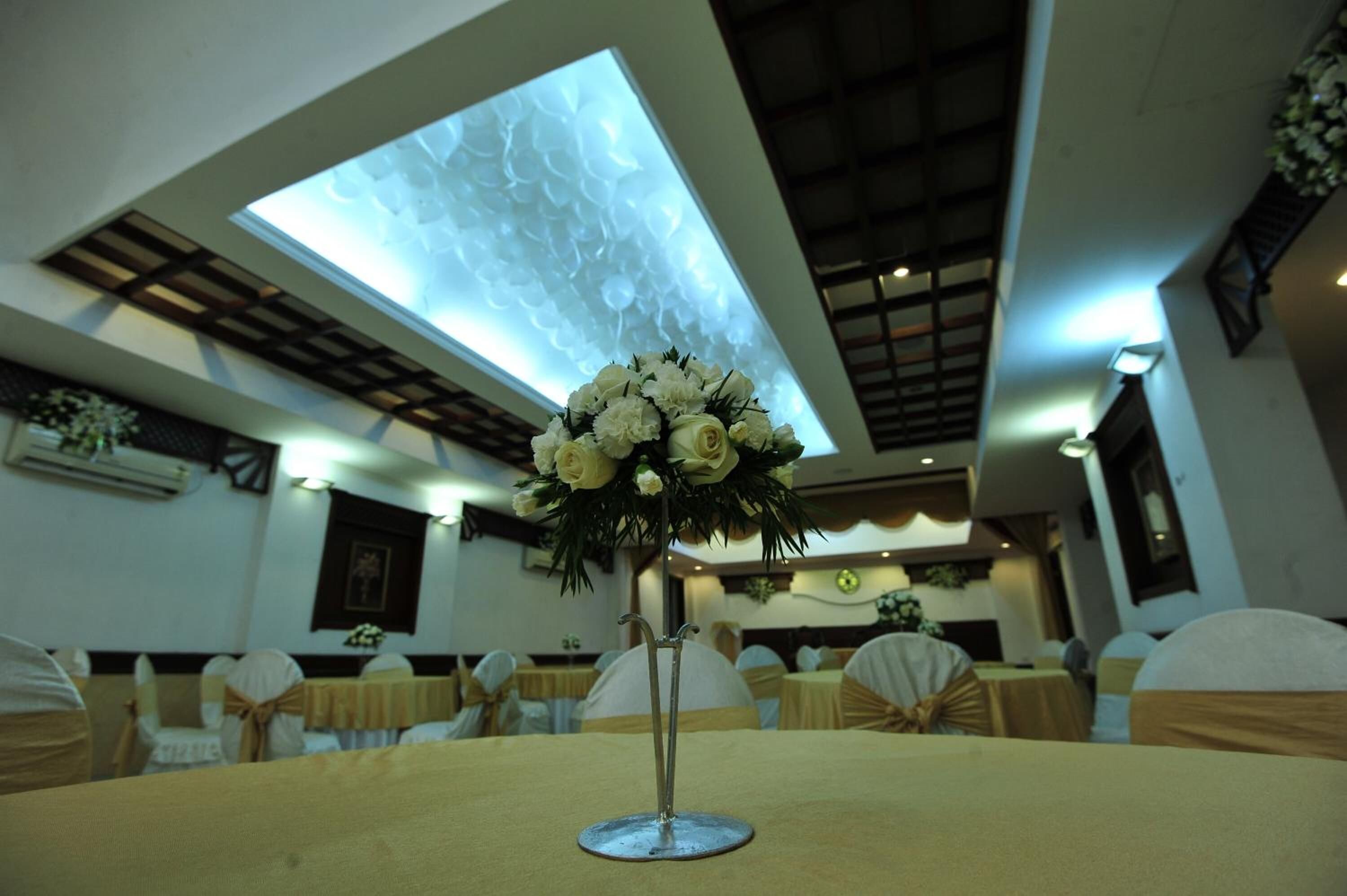 Ein Festsaal mit runden Tischen, die mit beigen Tischdecken bedeckt sind, weißen Stühlen mit goldenen Bändern und einer floralen Dekoration auf jedem Tisch.