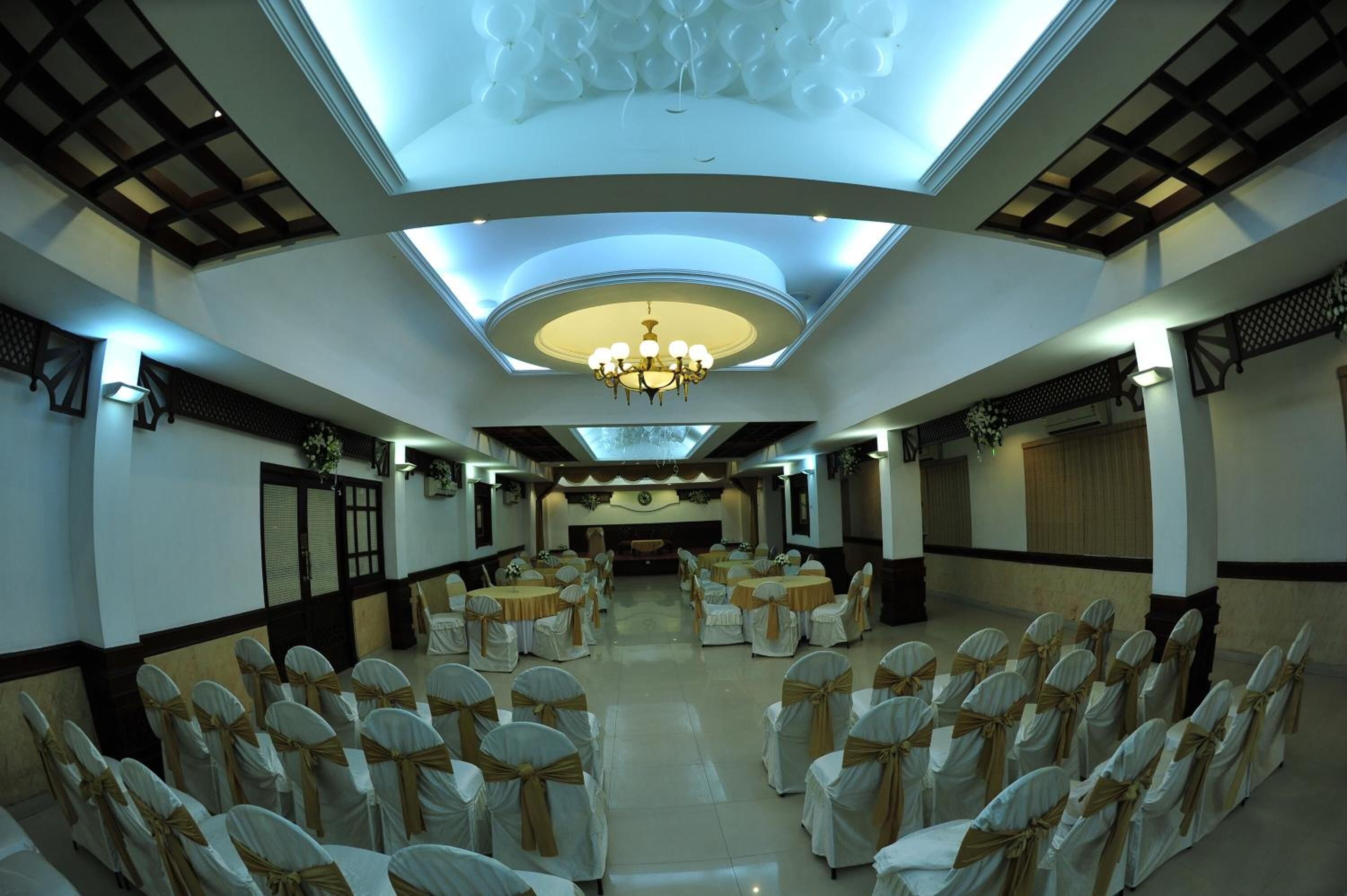 A banquet hall with rows of tables and chairs, a central chandelier, and decorative ceiling panels.