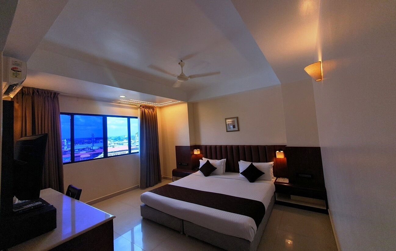 A hotel room with a large bed, a desk, a TV, a ceiling fan, and a window with a city view.