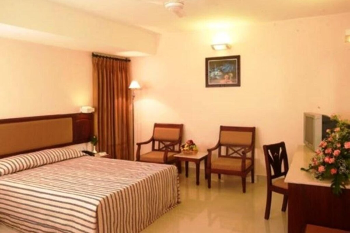 A hotel room with a bed, two chairs, a small table, a desk, a lamp, a picture on the wall, and a ceiling fan.