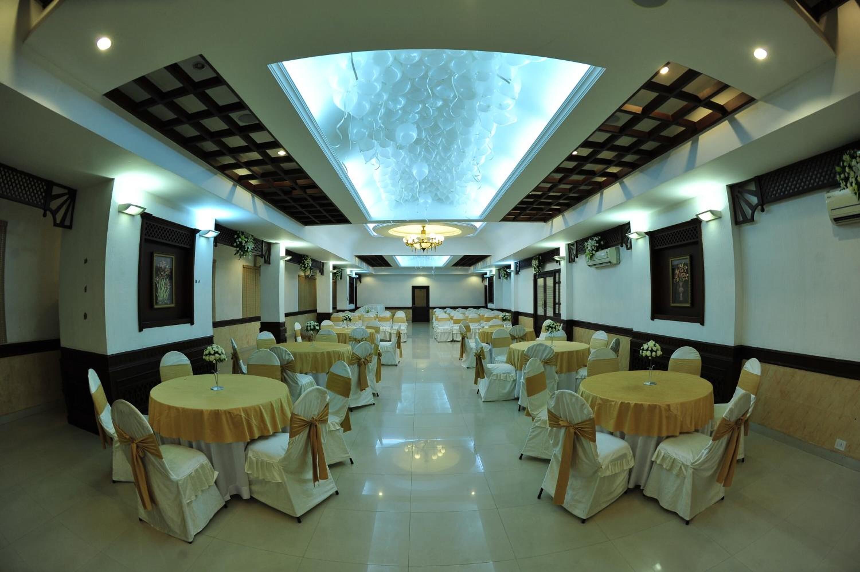 A banquet hall with round tables and chairs, a central chandelier, and a patterned ceiling.