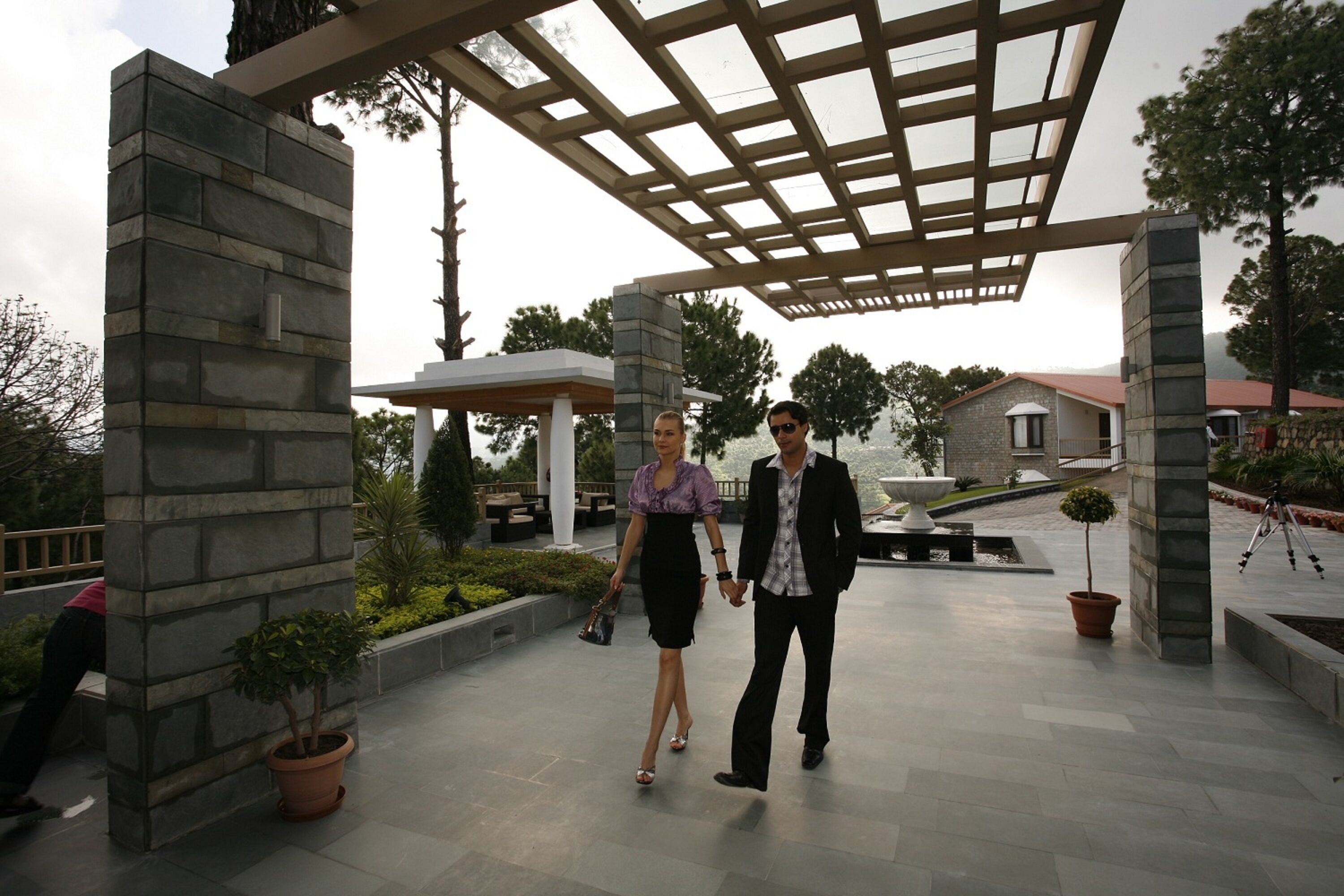 A covered walkway with stone pillars, a small fountain, and potted plants.