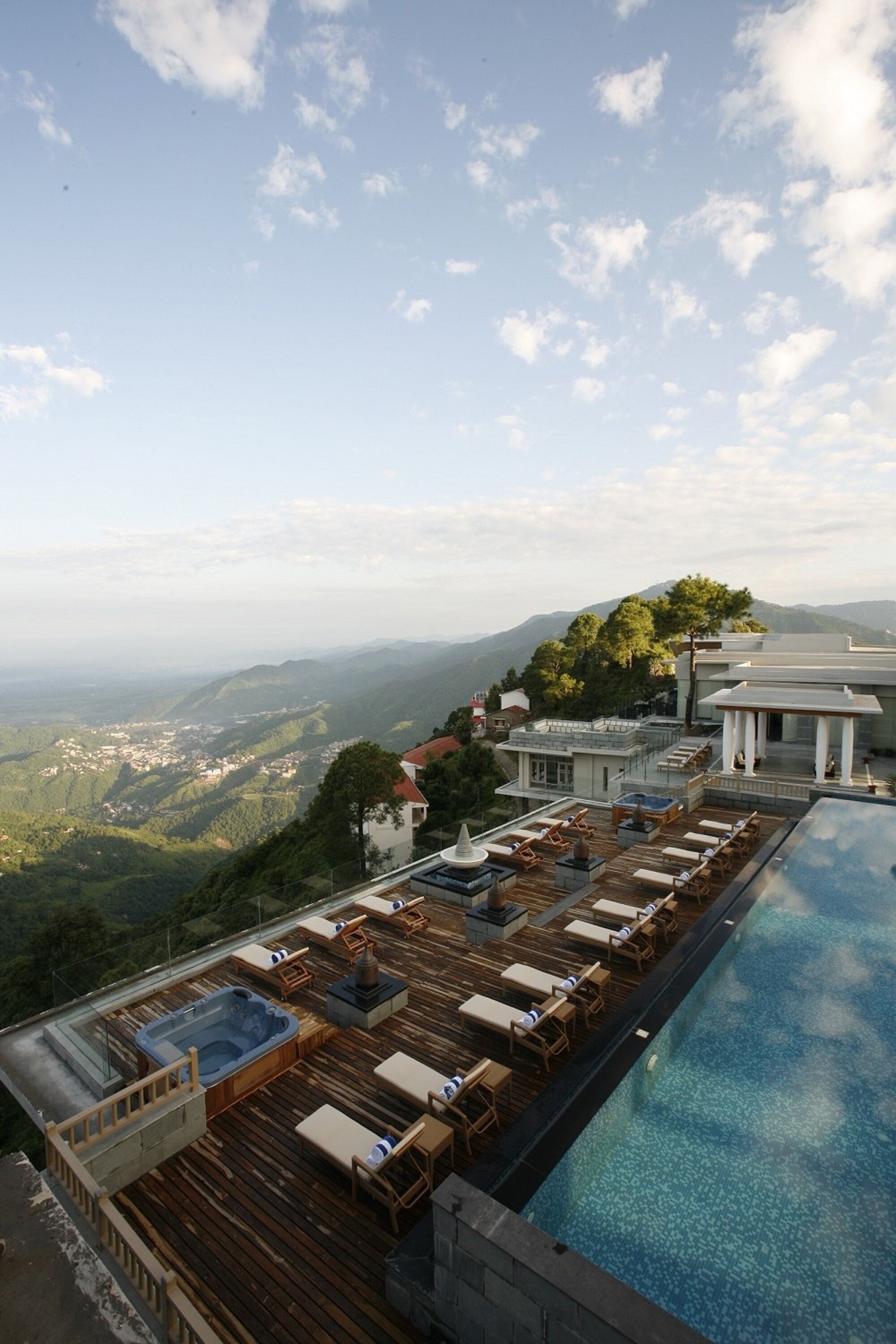 A rooftop infinity pool with lounge chairs and a hot tub, overlooking a mountainous landscape.