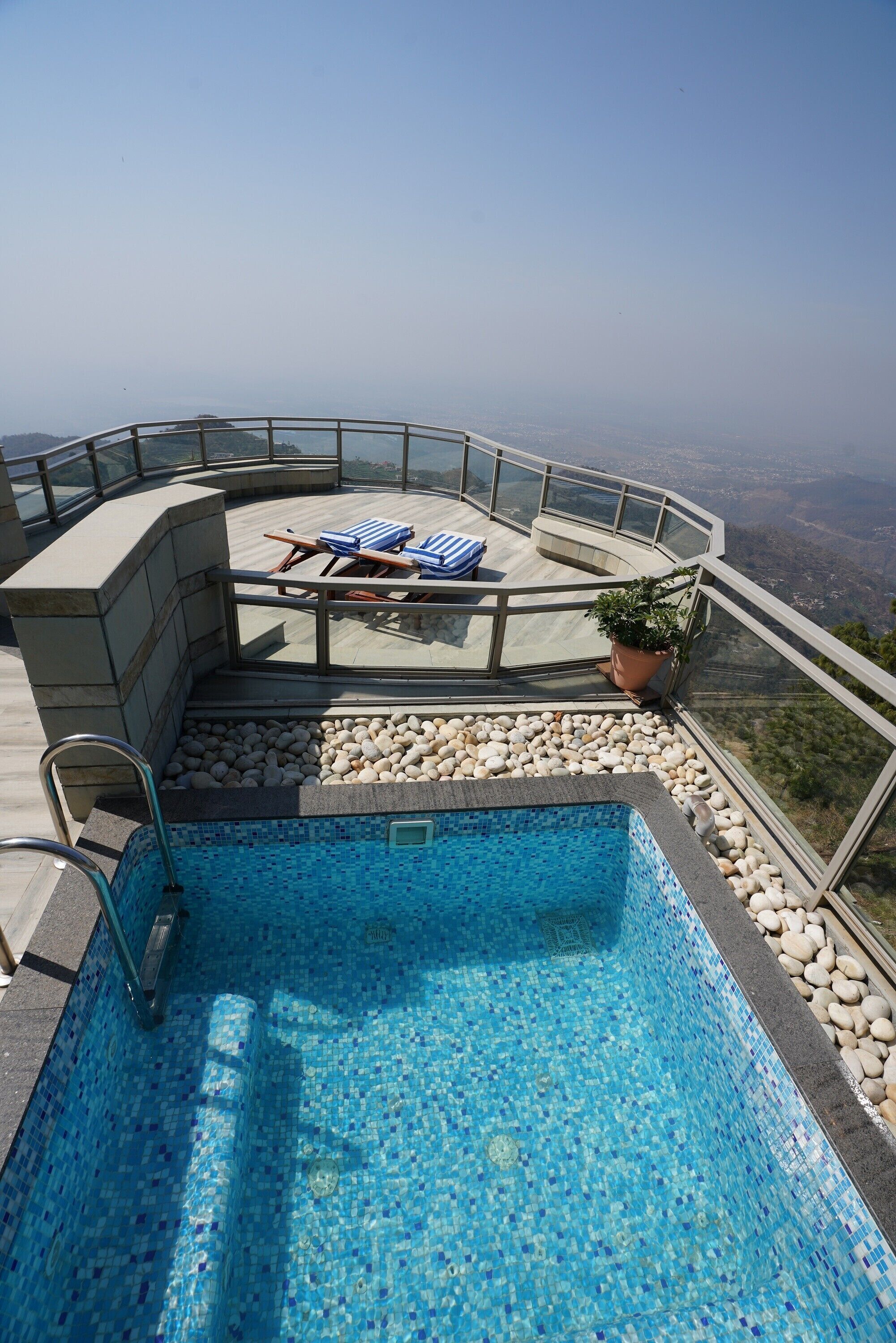 A rooftop pool with a clear view of the landscape, a seating area with lounge chairs, and a potted plant.