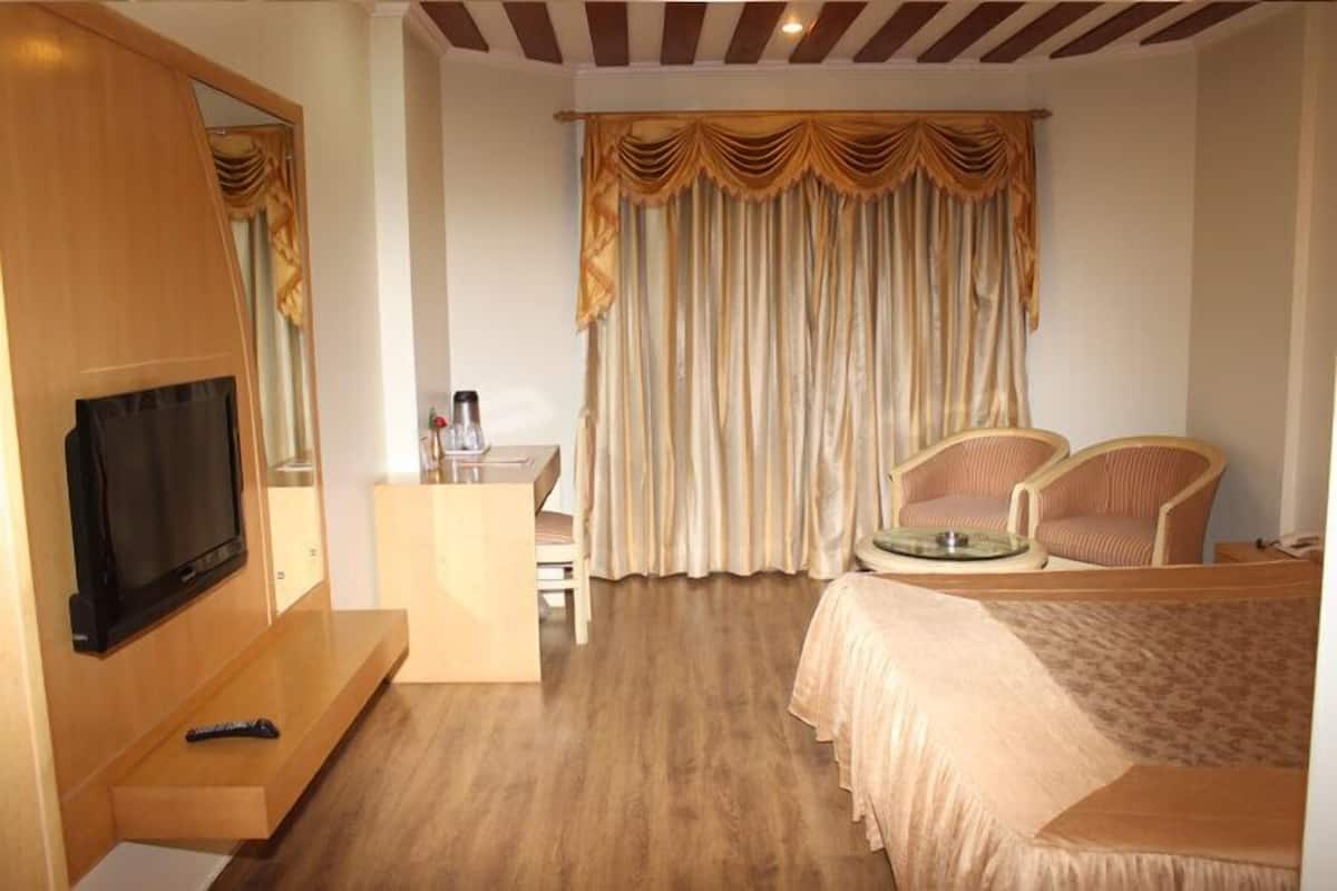 A hotel room with a TV, a desk, two chairs, a bed, and a large window with curtains.