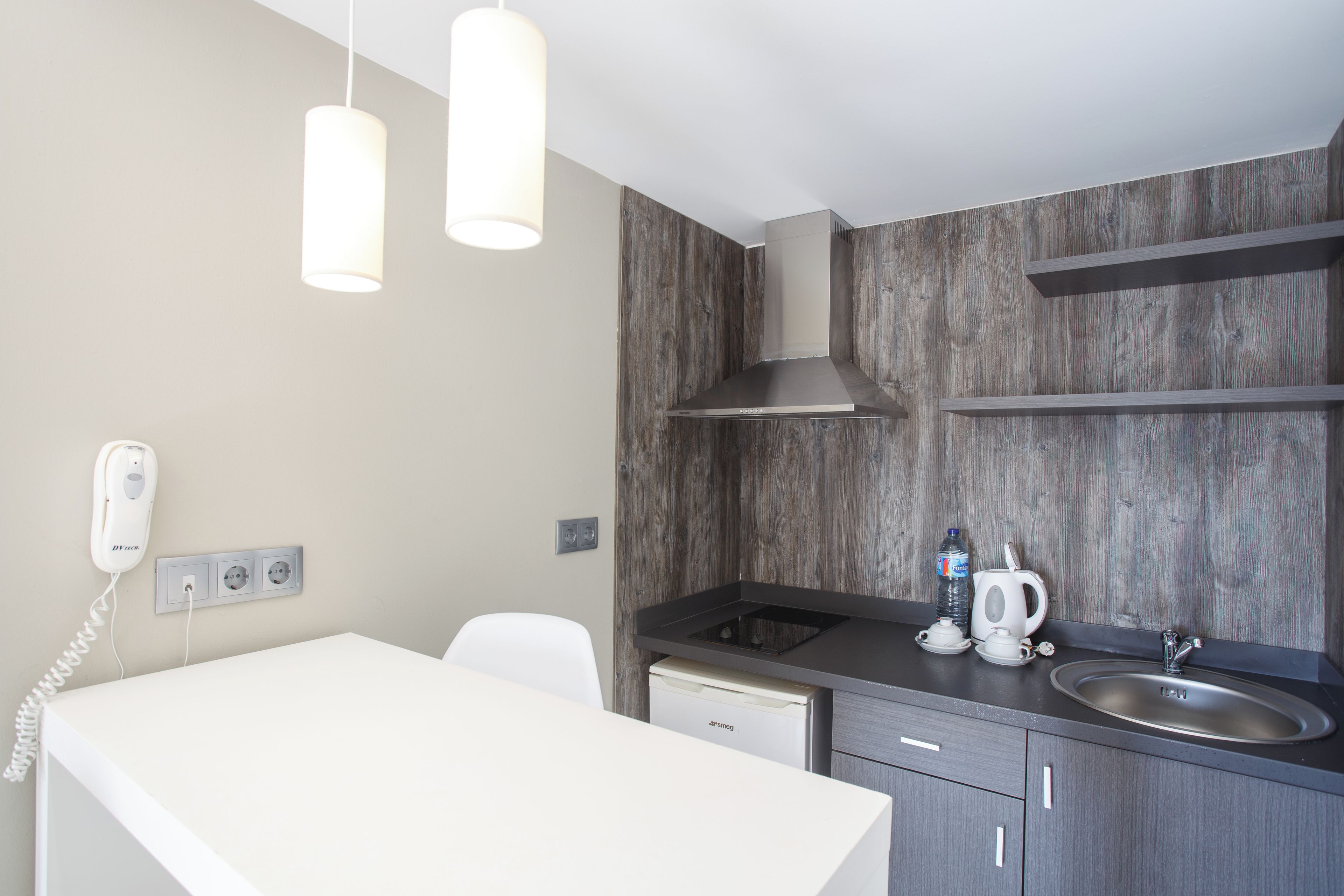 Una cocina moderna con encimera blanca, pileta de acero inoxidable y estufa integrada. Hay luces colgantes sobre la encimera y un teléfono instalado en la pared.
