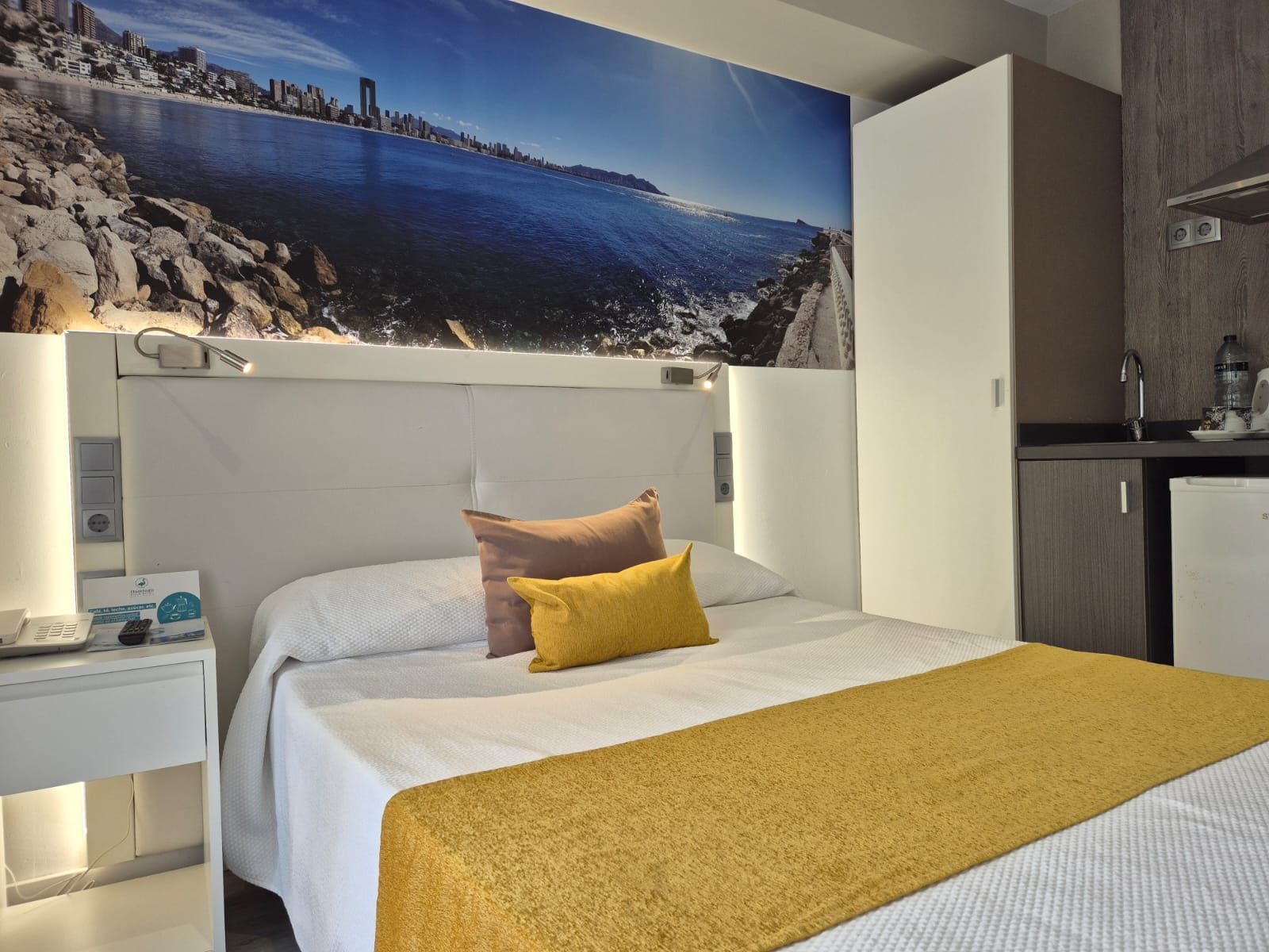 A hotel room with a bed, a nightstand, a wall-mounted TV, and a view of a coastal cityscape.