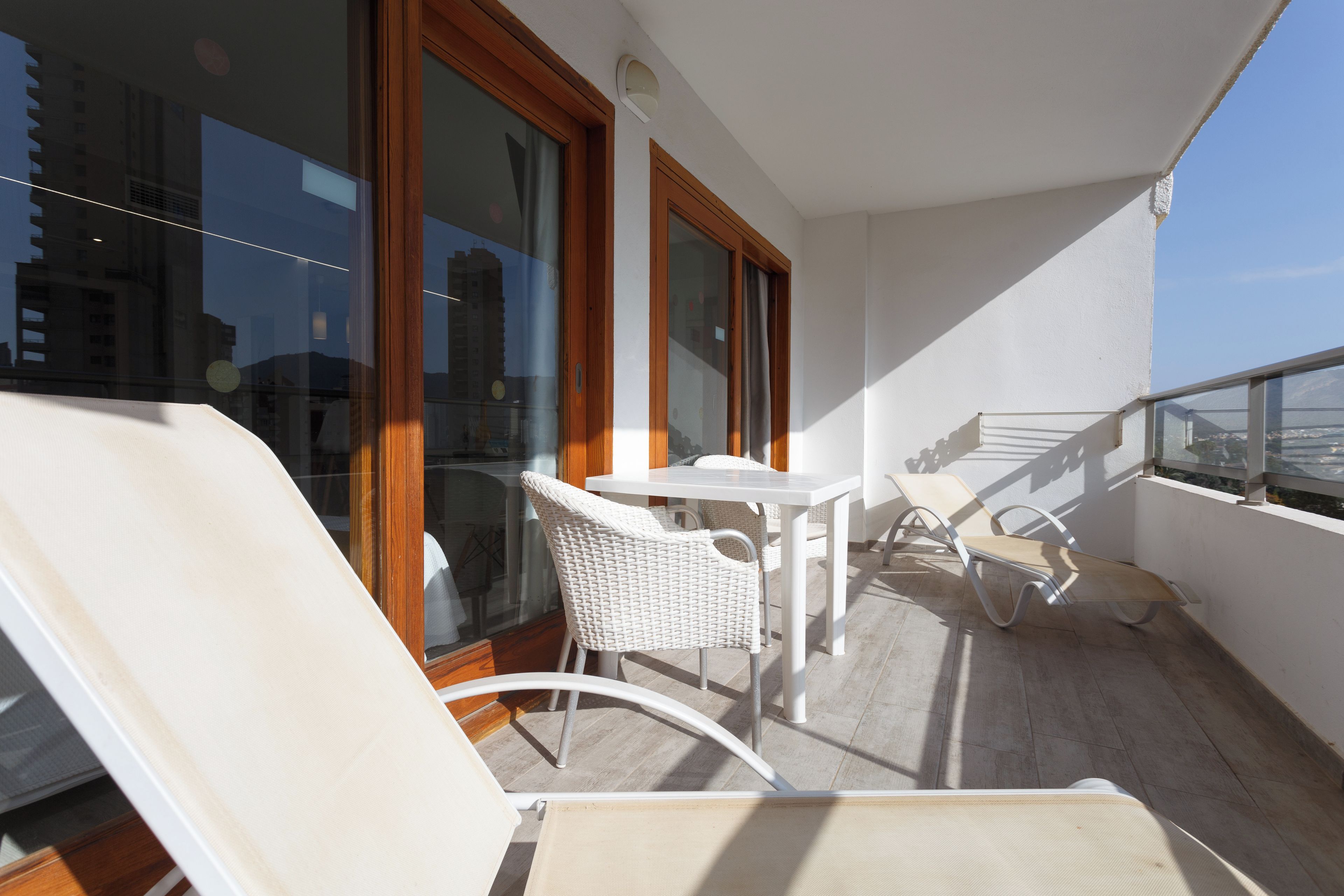 Ein Balkon mit weißen Korbmöbeln, darunter ein Tisch und zwei Stühle, mit Blick auf eine Stadtlandschaft.