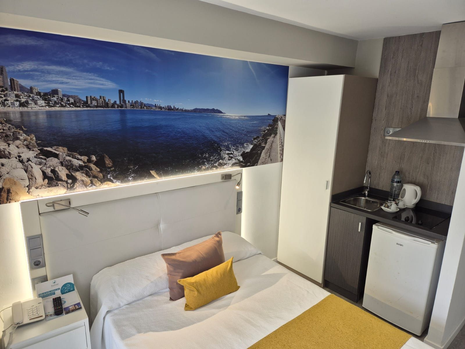 A hotel room with a bed, a view of a coastal cityscape, a kitchenette, and a small refrigerator.