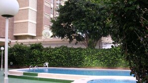 Zona de piscina con una piscina pequeña y una más grande, rodeada de follaje y un edificio al fondo.