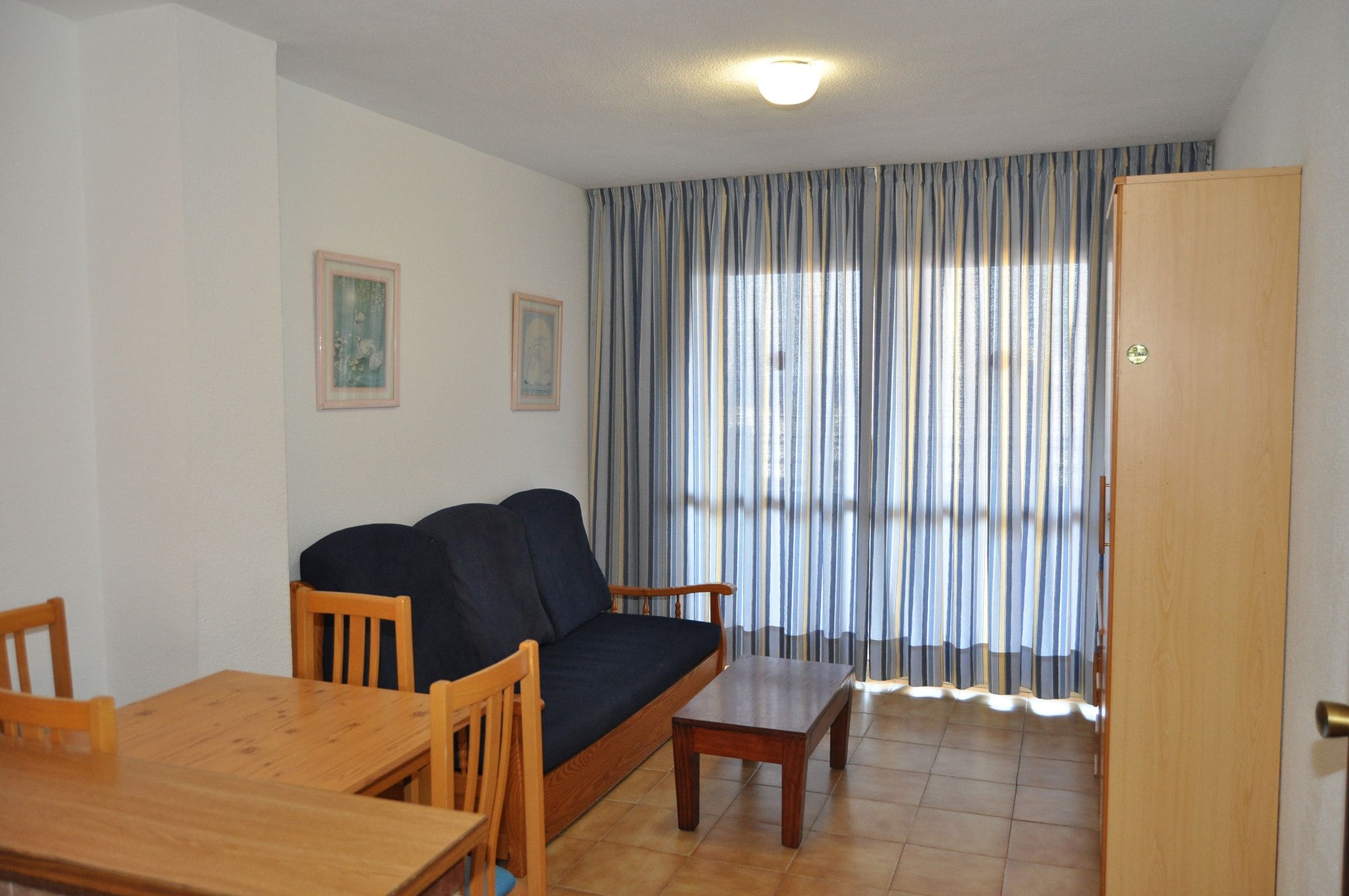 Una sala de estar con sofá, mesa de madera, sillas y un amplio ventanal con cortinas.
