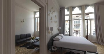 A hotel room with a bed, a sofa, a small table with a vase, and large windows with curtains.