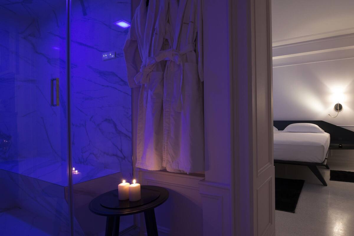 A modern hotel room with a marble bathroom, a bedside table with candles, and a bed with a black headboard.