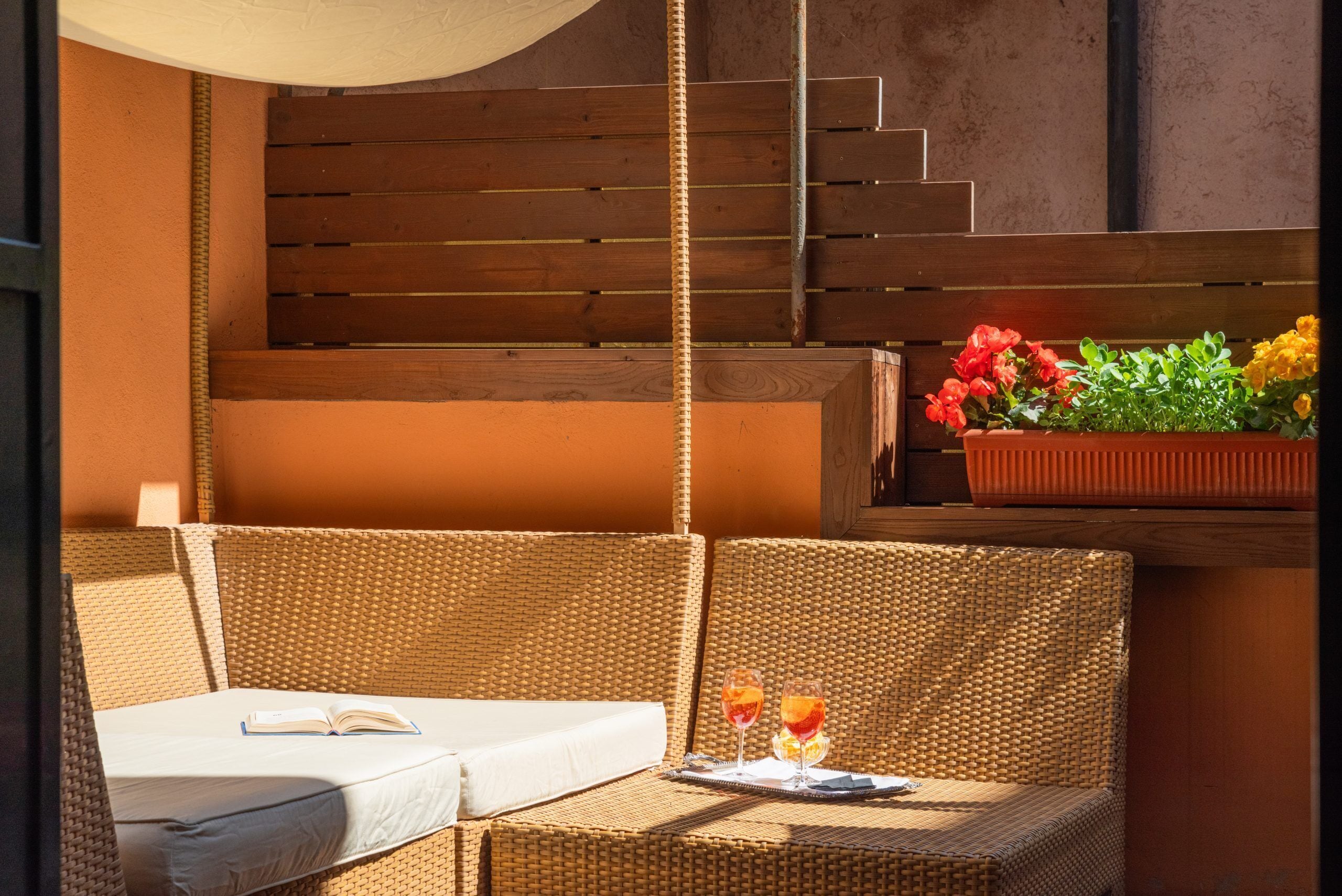 A wicker lounge chair with a white cushion and an open book, two glasses with drinks, and a planter with flowers.