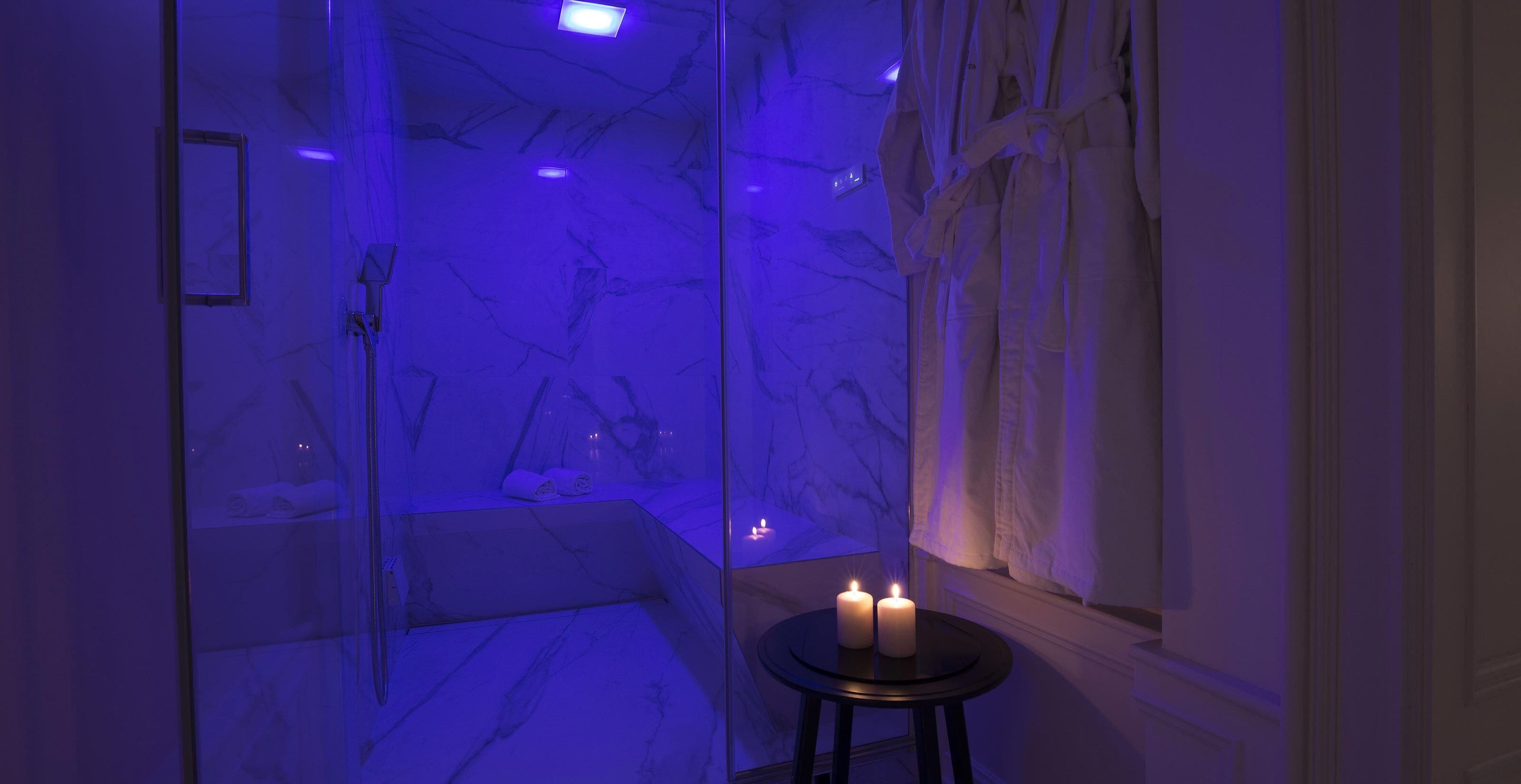 Un baño con cabina de ducha de vidrio, paredes de mármol y dos velas encendidas sobre un taburete.