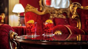Duas taças de bebida vermelha com fatias de laranja sobre uma mesa num quarto com estofados de veludo vermelho e mobiliário dourado detalhado.