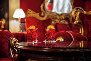 Duas taças de bebida vermelha com fatias de laranja sobre uma mesa num quarto com estofados de veludo vermelho e mobiliário dourado detalhado.