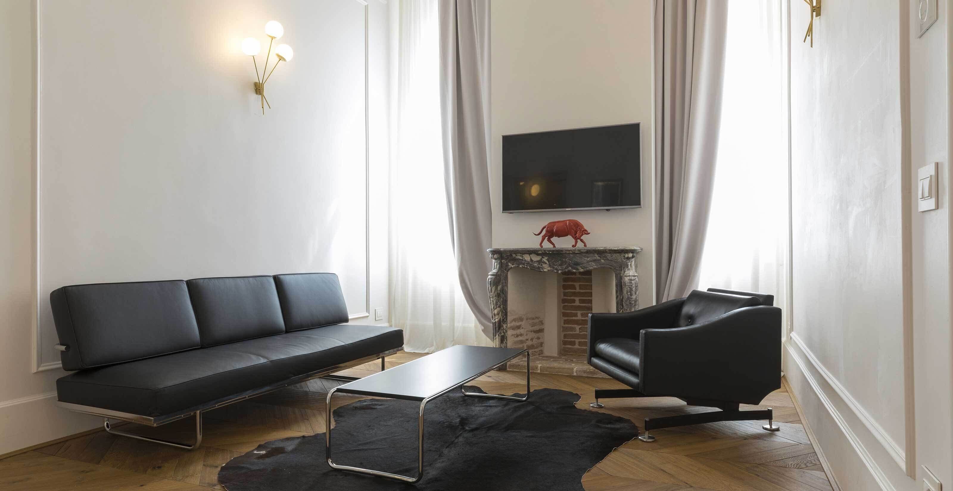 Ein modernes Wohnzimmer mit einer schwarzen Couch, einem schwarzen Sessel, einem kleinen Tisch, einem Kamin mit einem roten Dekostück und einem an der Wand befestigten Flachbildfernseher.