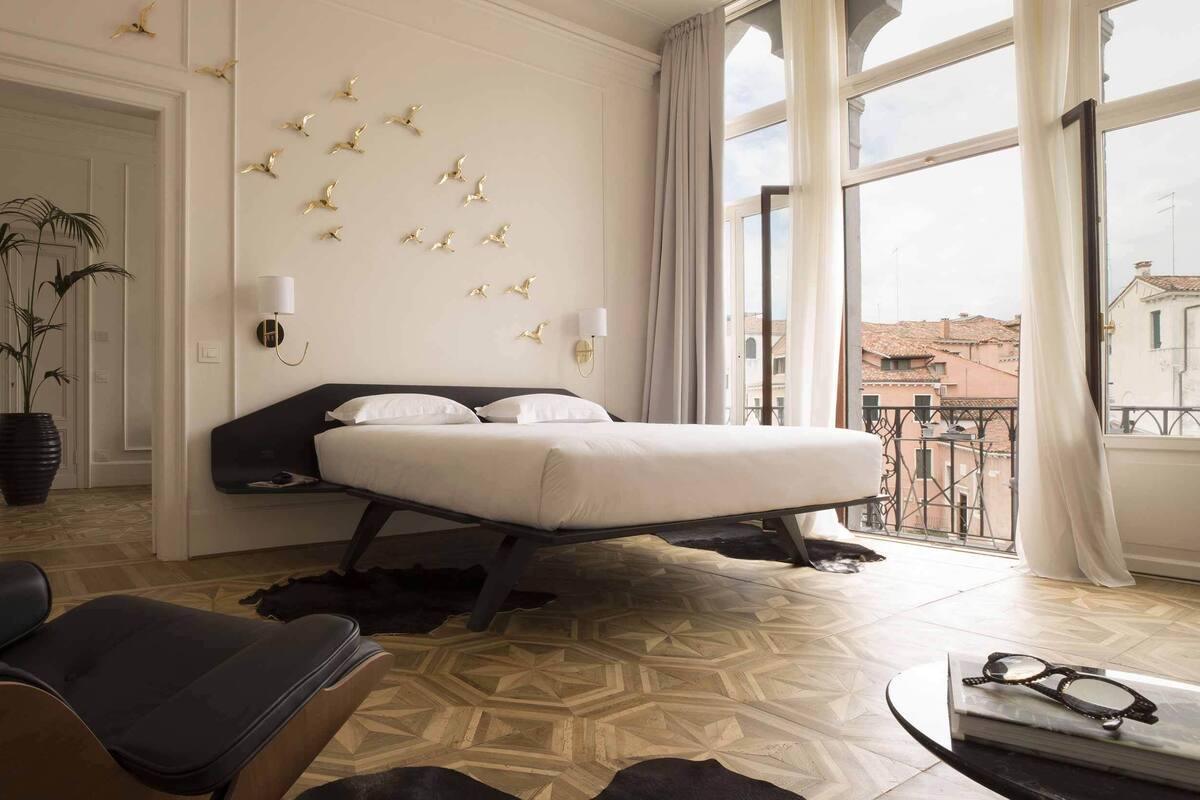 A hotel room with a bed, a chair, a potted plant, a wall-mounted lamp, and a view of buildings through large windows.