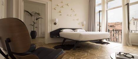 Hotelkamer met een bed, een stoel, een potplant, een wandlamp en uitzicht op gebouwen door grote ramen.
