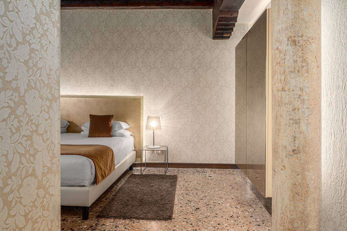 A bedroom with a bed, a nightstand, a lamp, a patterned wall, and a textured floor.