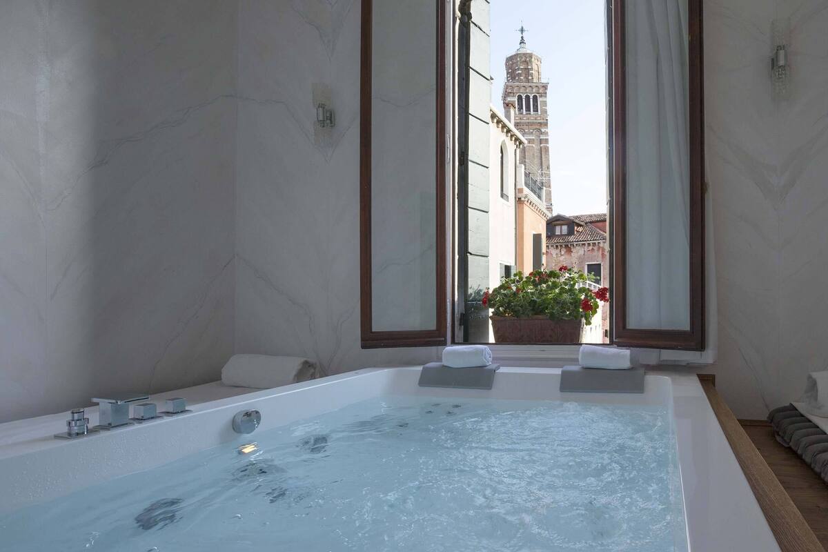A modern bathroom with a large bathtub, a window with a view of a building, and a potted plant.