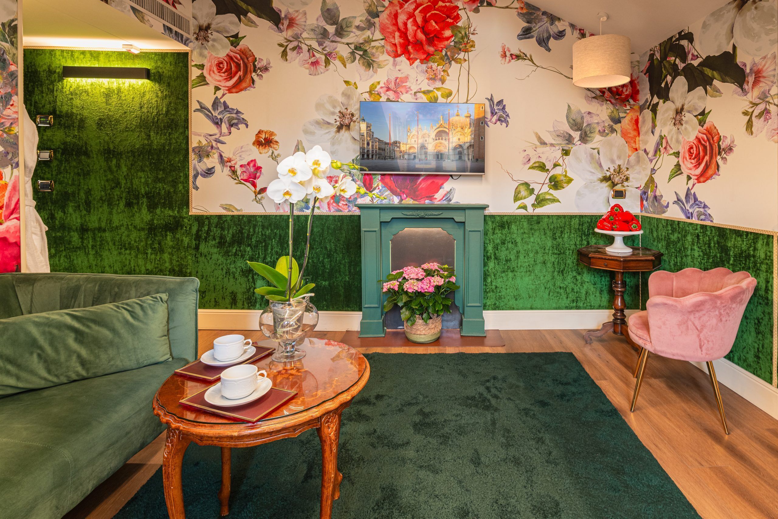 A room with a green textured wall, a fireplace, a sofa, a table with cups and saucers, a pink chair, and a floral mural.