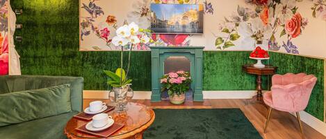 A room with a green textured wall, a fireplace, a sofa, a table with cups and saucers, a pink chair, and a floral mural.