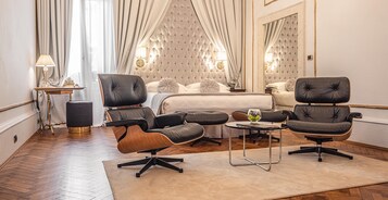 Una habitación de hotel con una cama, dos sillones de cuero, una mesita con un jarrón y un espejo grande.