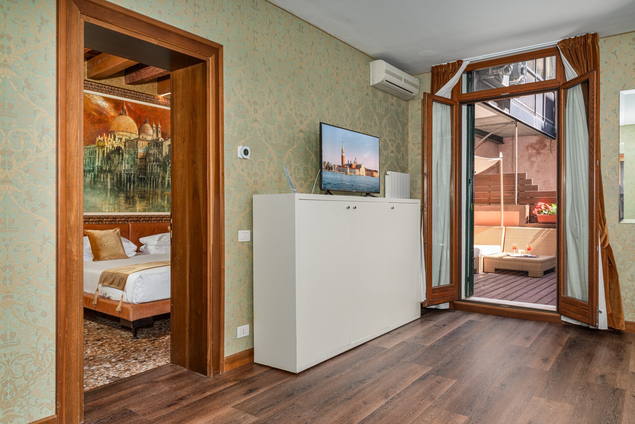 Quarto com cama, televisão em um armário, pintura de uma paisagem urbana e vista para uma área de estar ao ar livre.