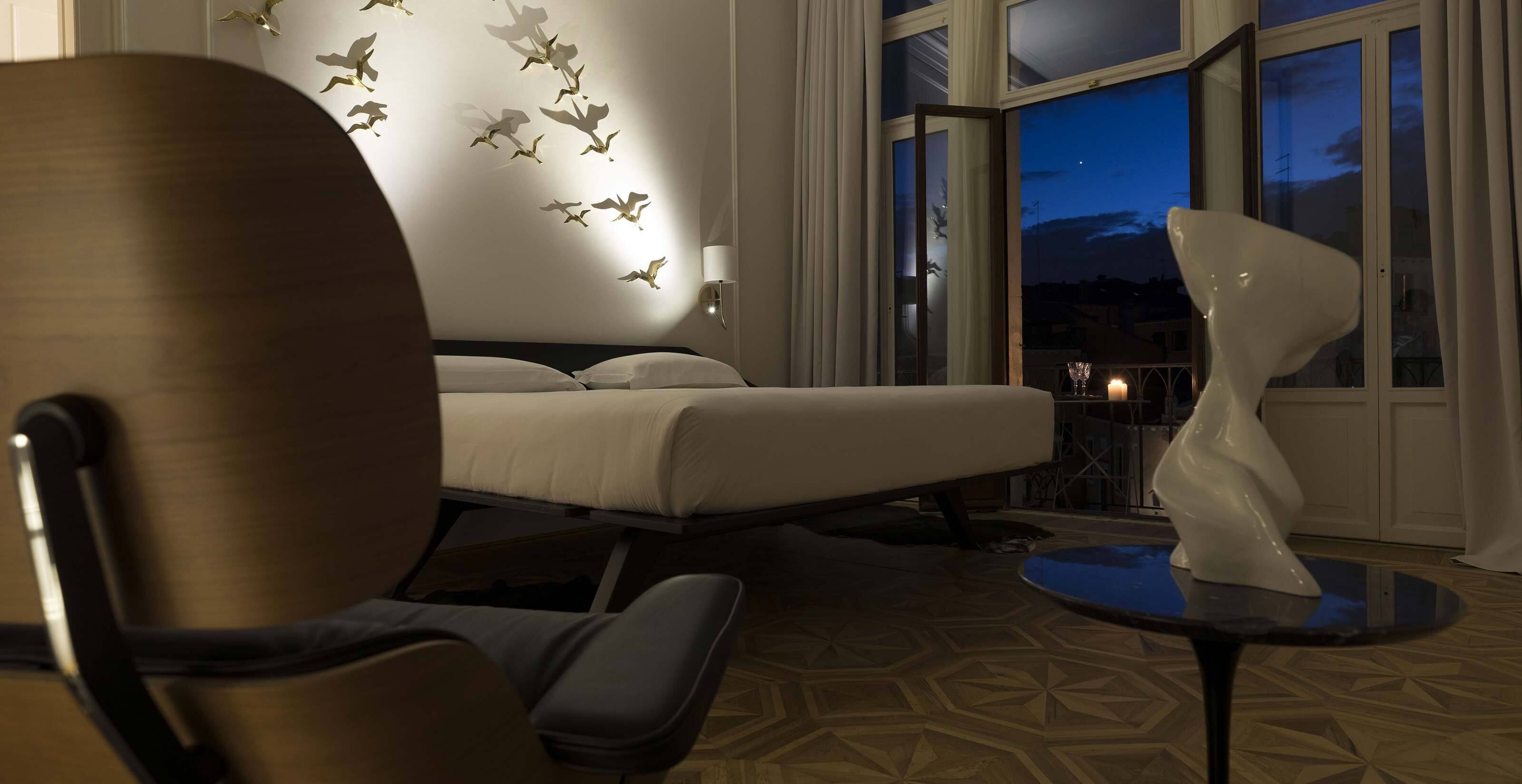 A hotel room with a bed, a chair, a table, a wall with bird decals, and a view of the night sky through large windows.