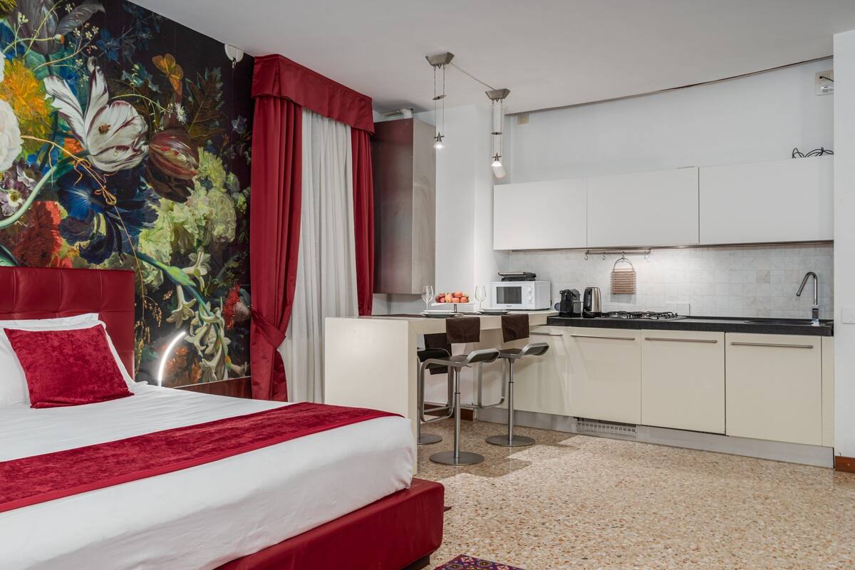 A hotel room with a bed, a red headboard, a large floral mural, a kitchenette, and a bar area.
