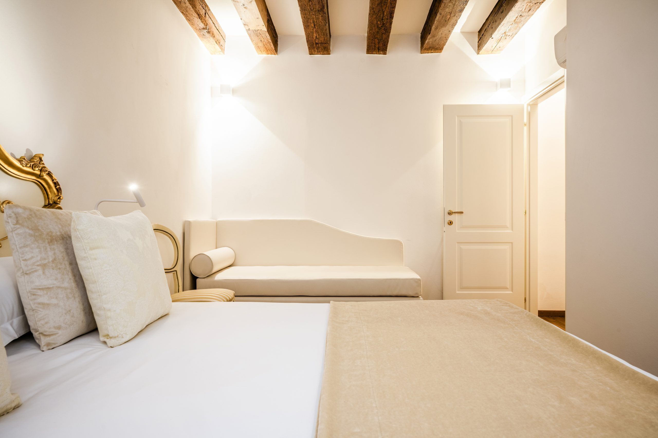 A bedroom with a bed, a bench, a door, and wooden beams on the ceiling.
