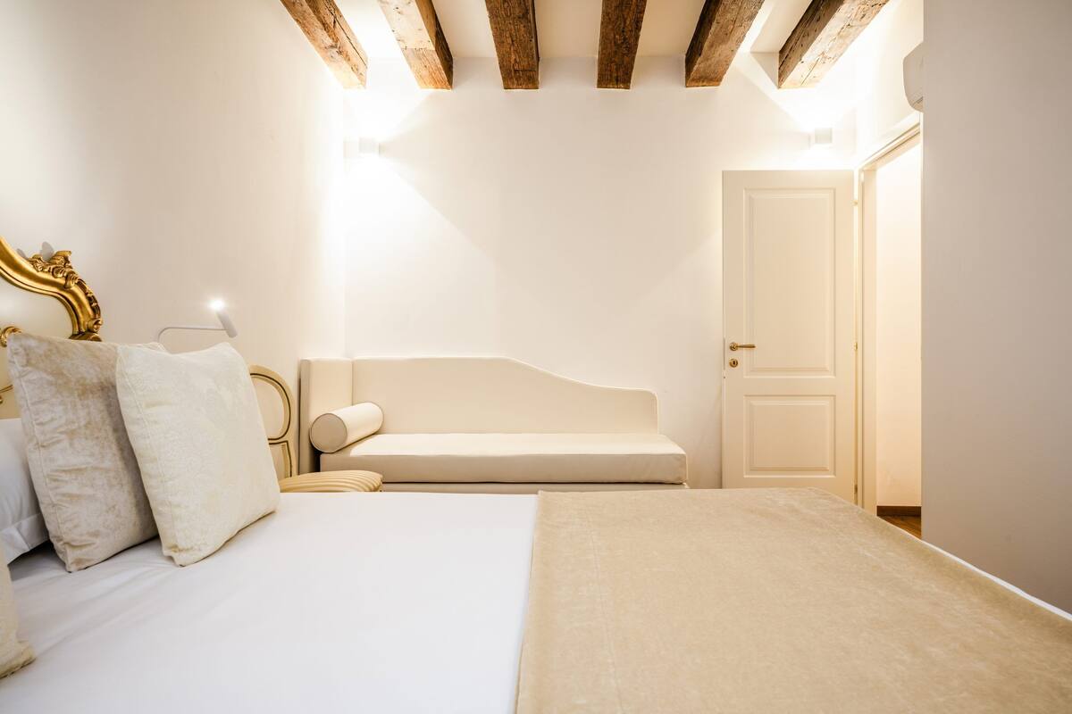 A bedroom with a bed, a bench, a door, and wooden beams on the ceiling.