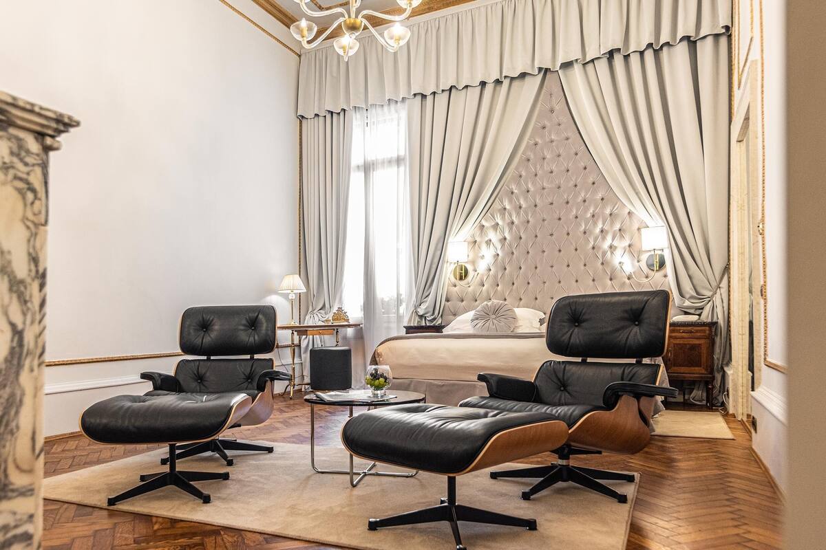 A hotel room with a bed, two black leather chairs, a small table with a lamp, and a large window with curtains.