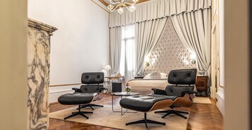Una habitación de hotel con una cama, dos sillones de cuero negro, una mesita de noche con una lámpara y un amplio ventanal con cortinas.