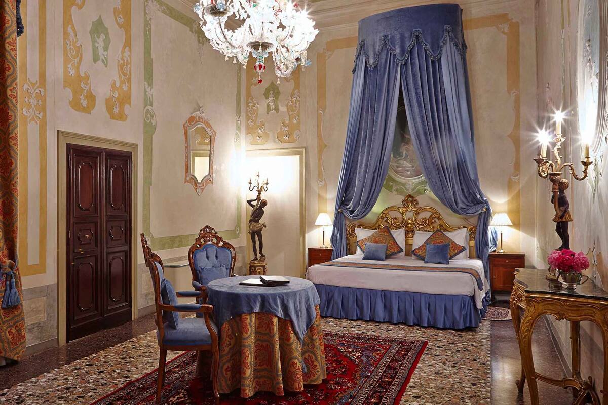 A luxurious bedroom with a four-poster bed, a chandelier, a round table, and a large mirror.