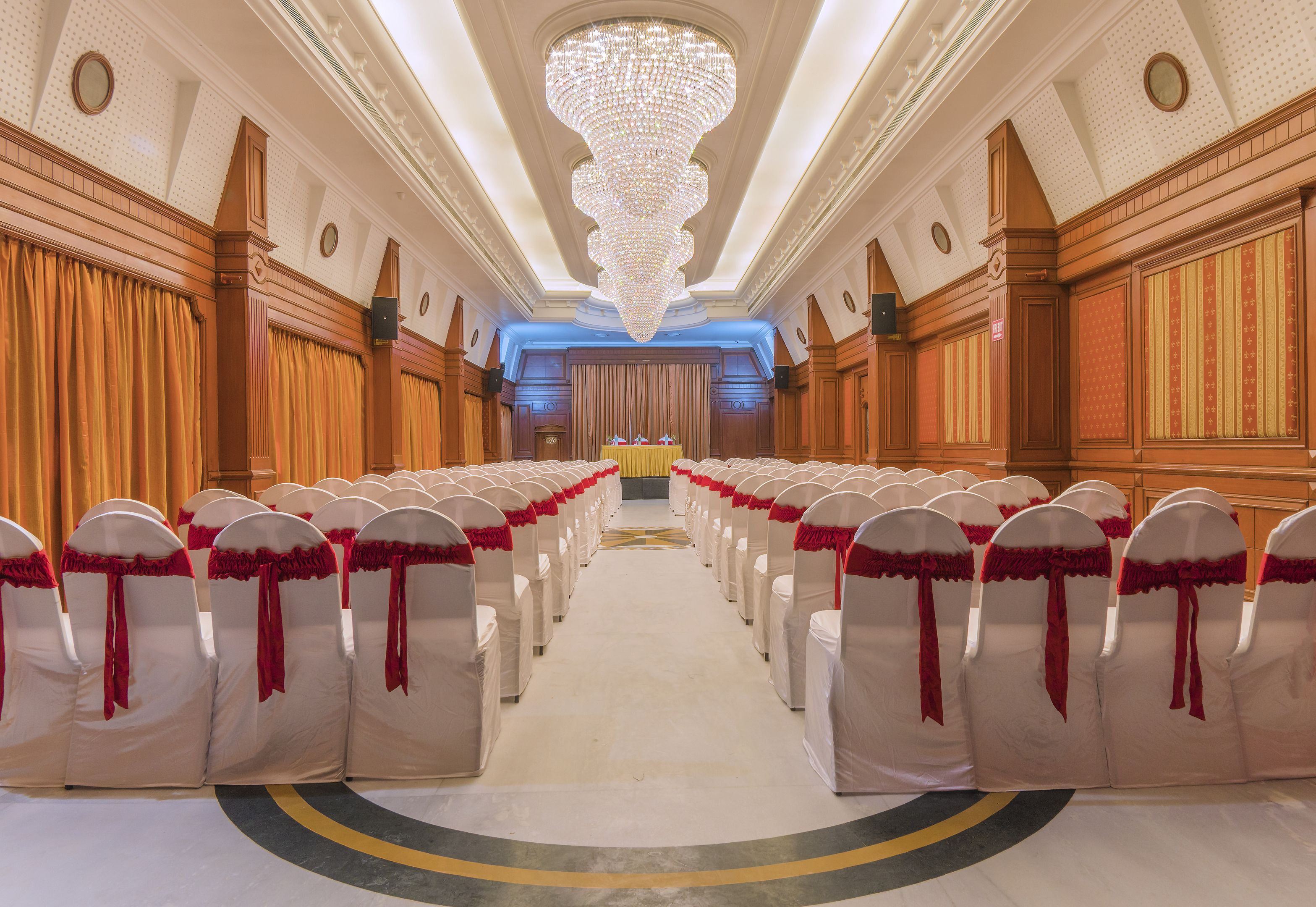 A hotel lobby with rows of white chairs with red sashes, a large chandelier, and wooden paneling on the walls.