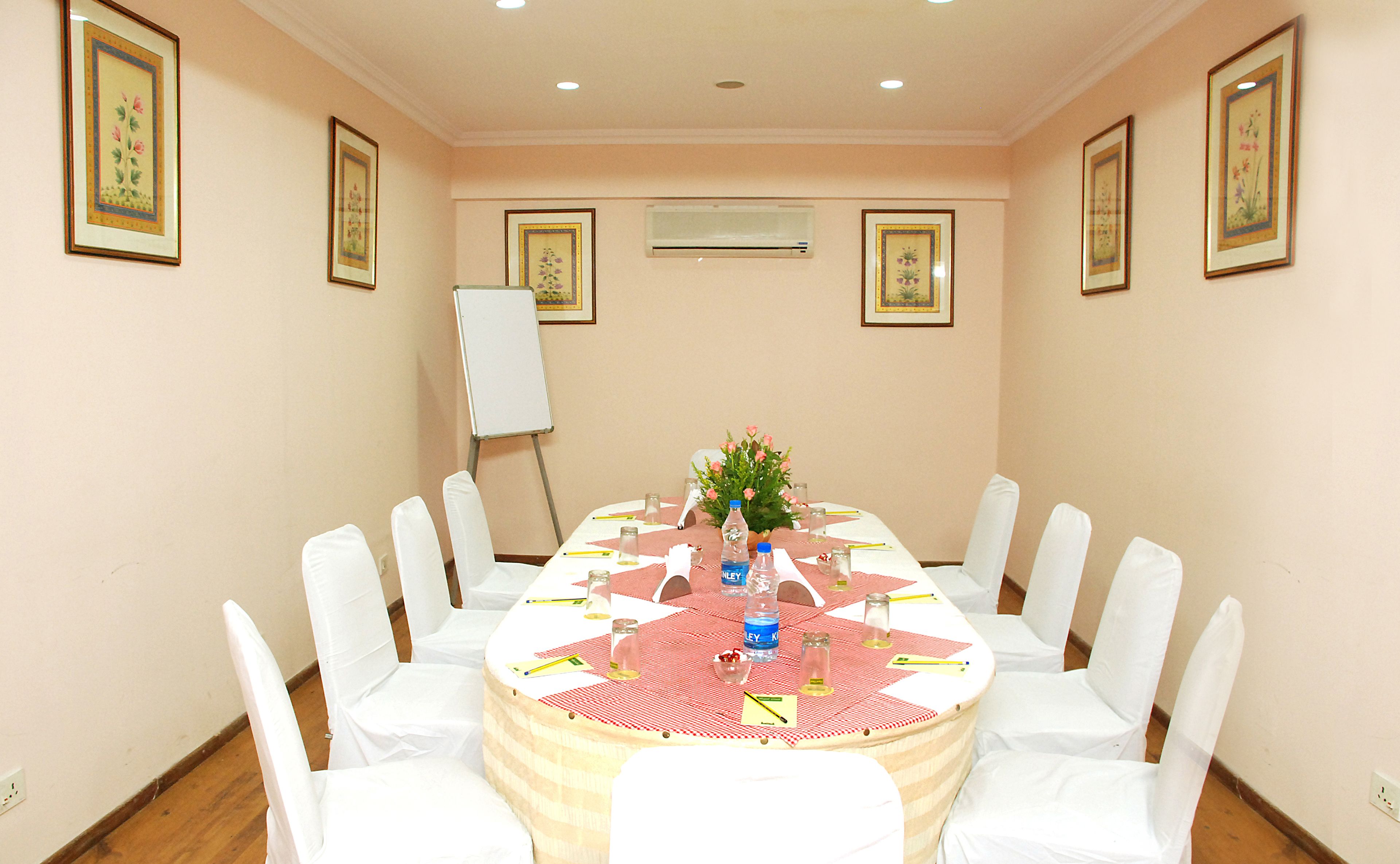 Un salón de conferencias con una mesa redonda, sillas blancas, una pizarra y cuadros con motivos florales en las paredes.