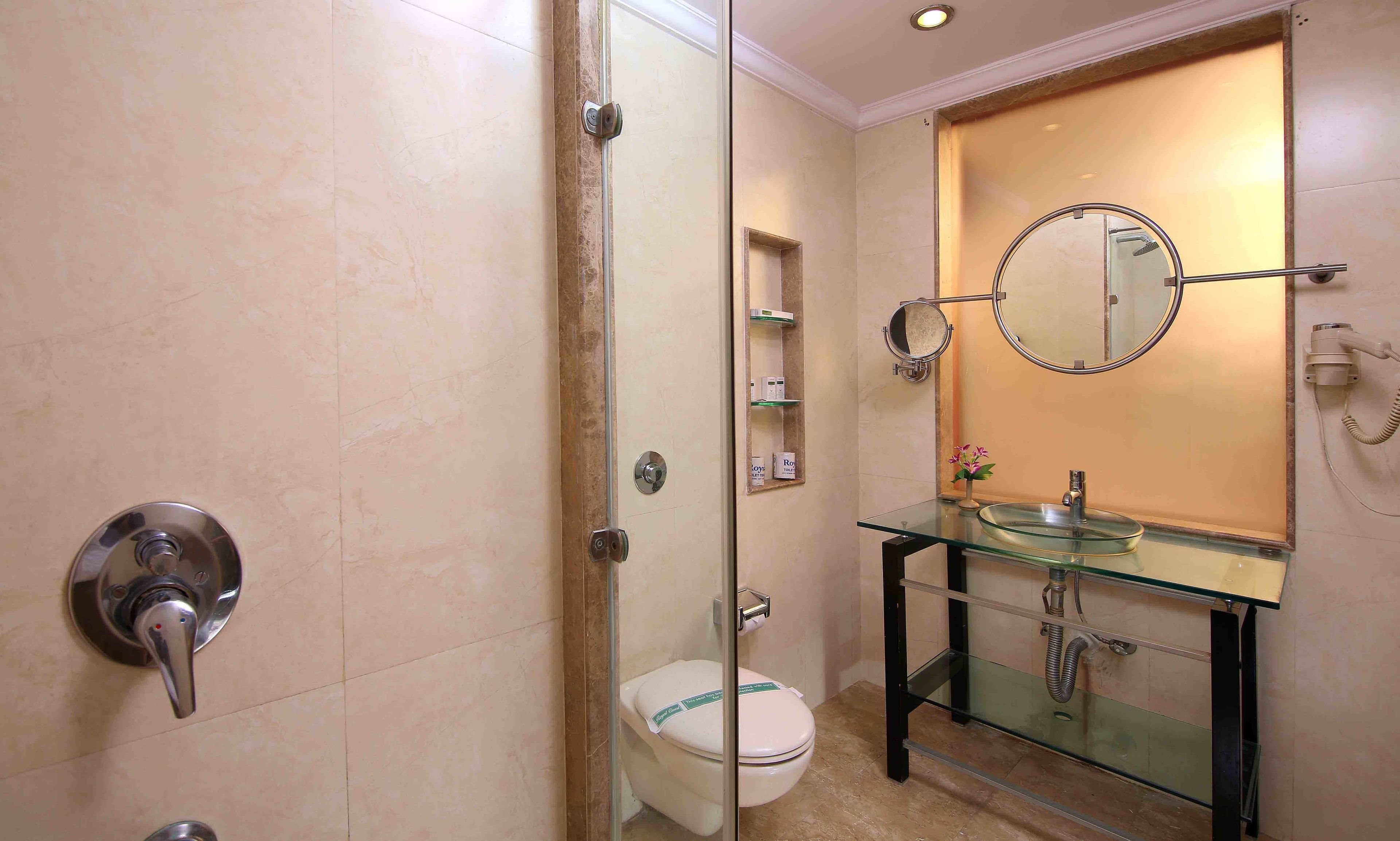 Un baño con ducha envuelta en vidrio, inodoro, lavamanos de vidrio con espejo y estante con artículos de aseo.