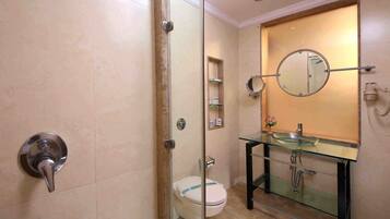Un baño con ducha envuelta en vidrio, inodoro, lavamanos de vidrio con espejo y estante con artículos de aseo.