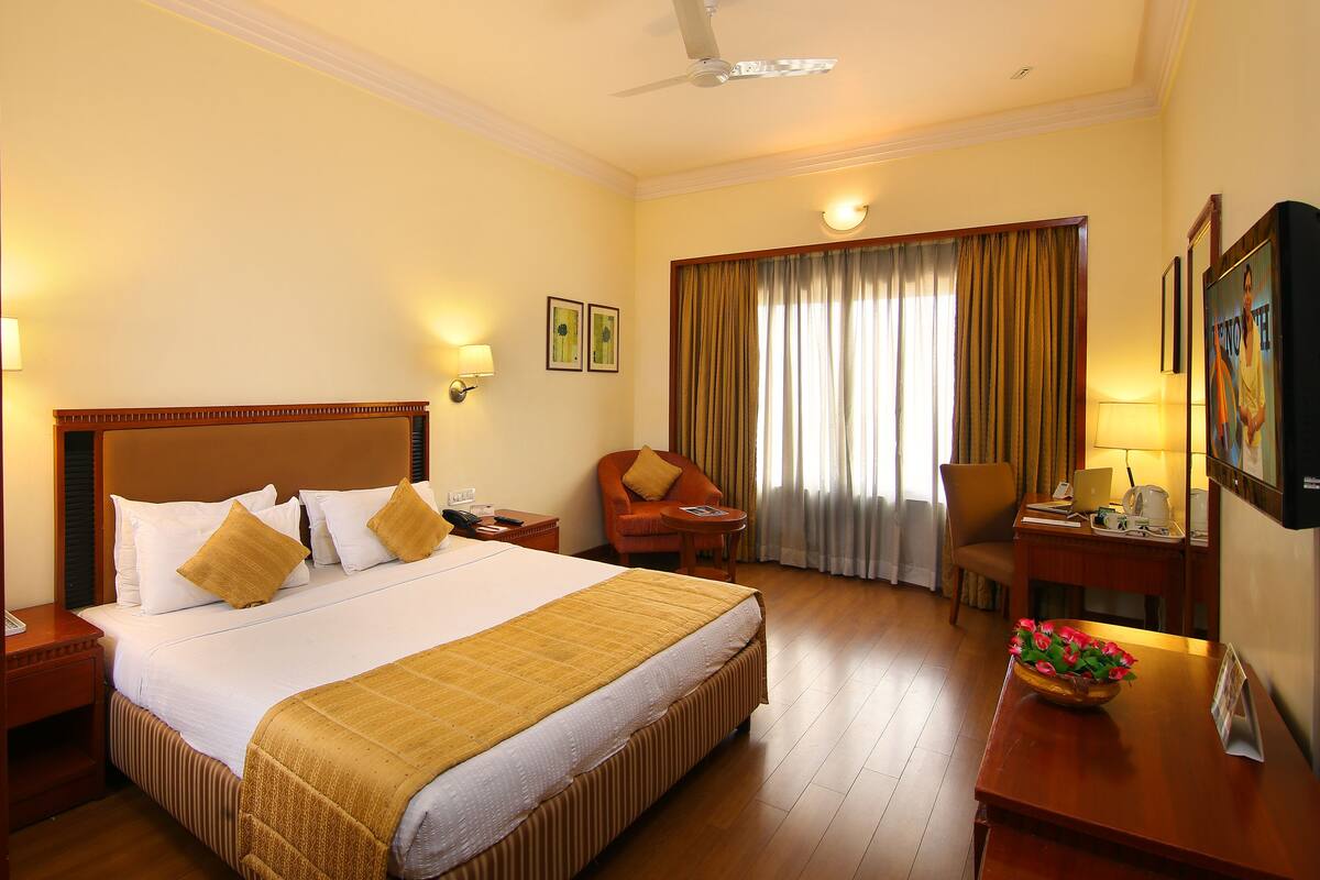 A hotel room with a large bed, a desk with a laptop, a chair, a desk lamp, and a window with curtains.